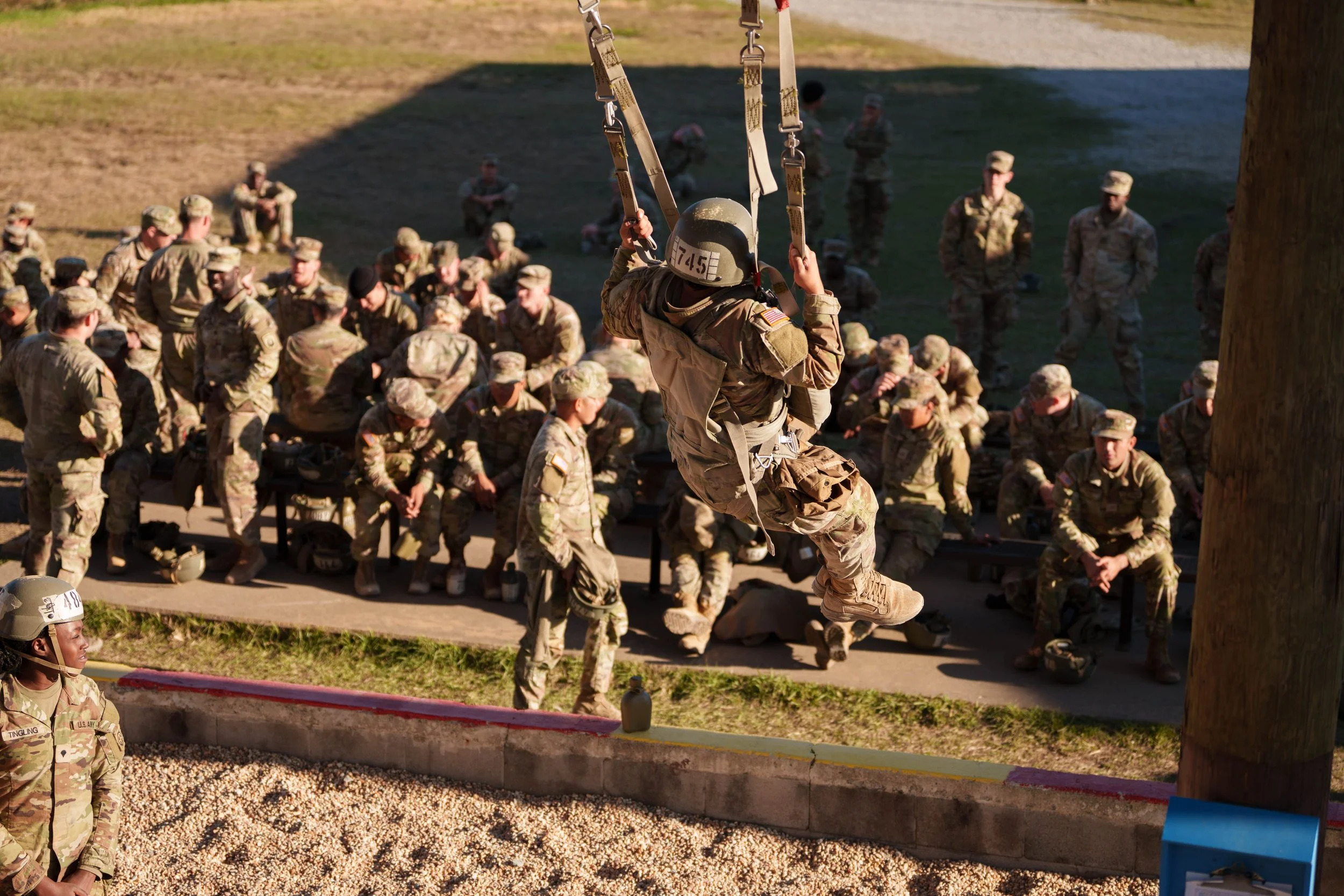 U.S. military personnel seated and standing outdoors, with one soldier in a harness jumping off to a lower level during training.