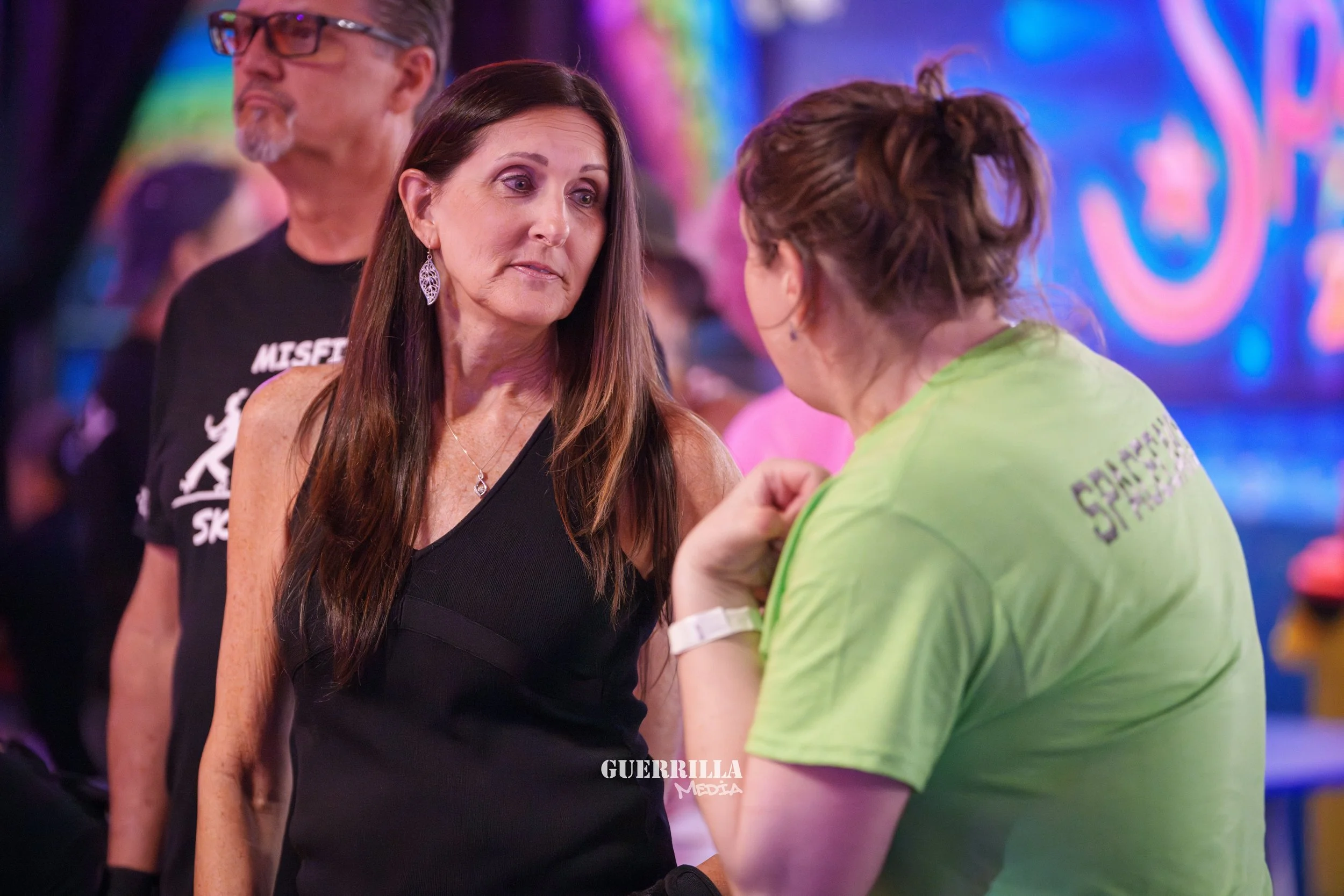 Two women are engaged in a serious conversation at an event, with colorful lights in the background. The woman on the left has long brown hair, wearing a black sleeveless top and jewelry. The woman on the right has curly hair tied back, wearing a bri