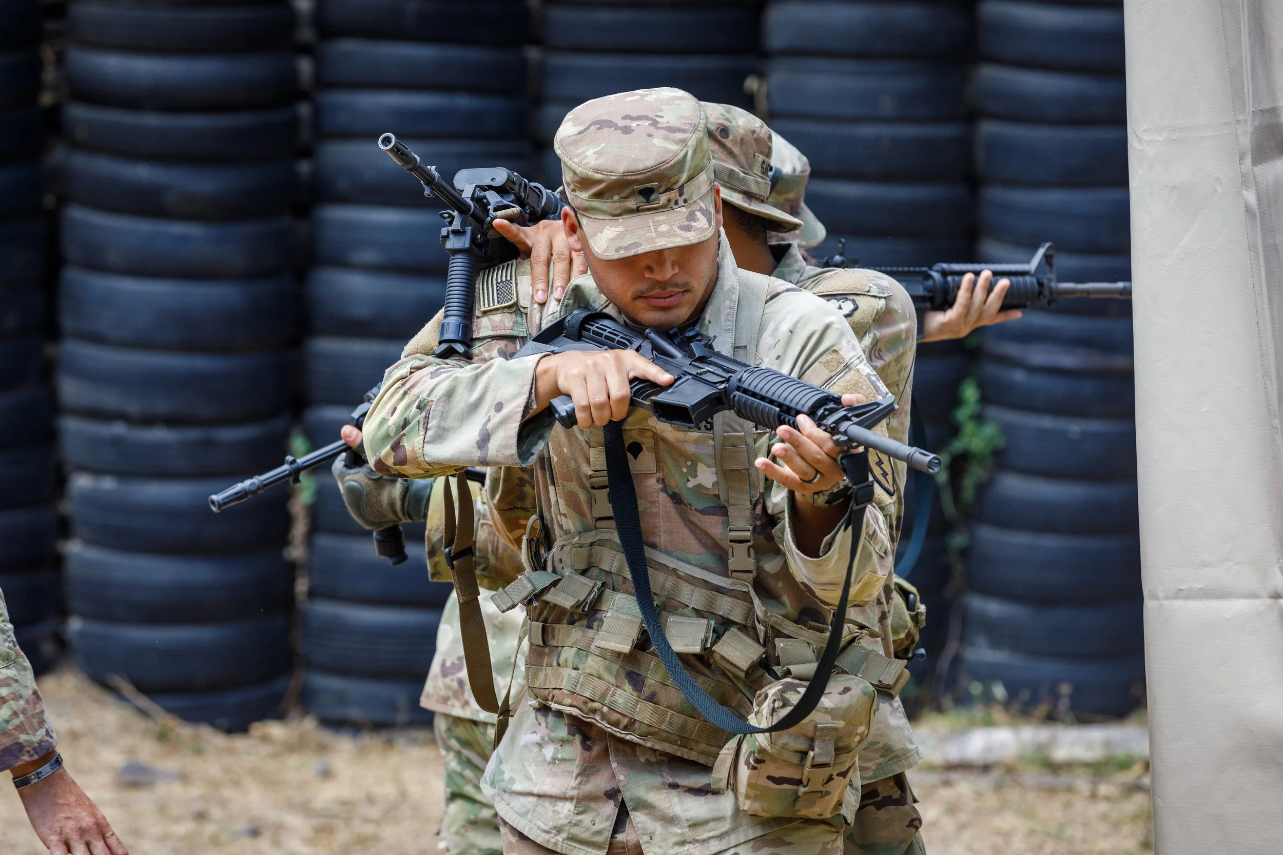 Soldiers in military camouflage uniforms practicing tactical shooting with rifles behind a barrier of tires.