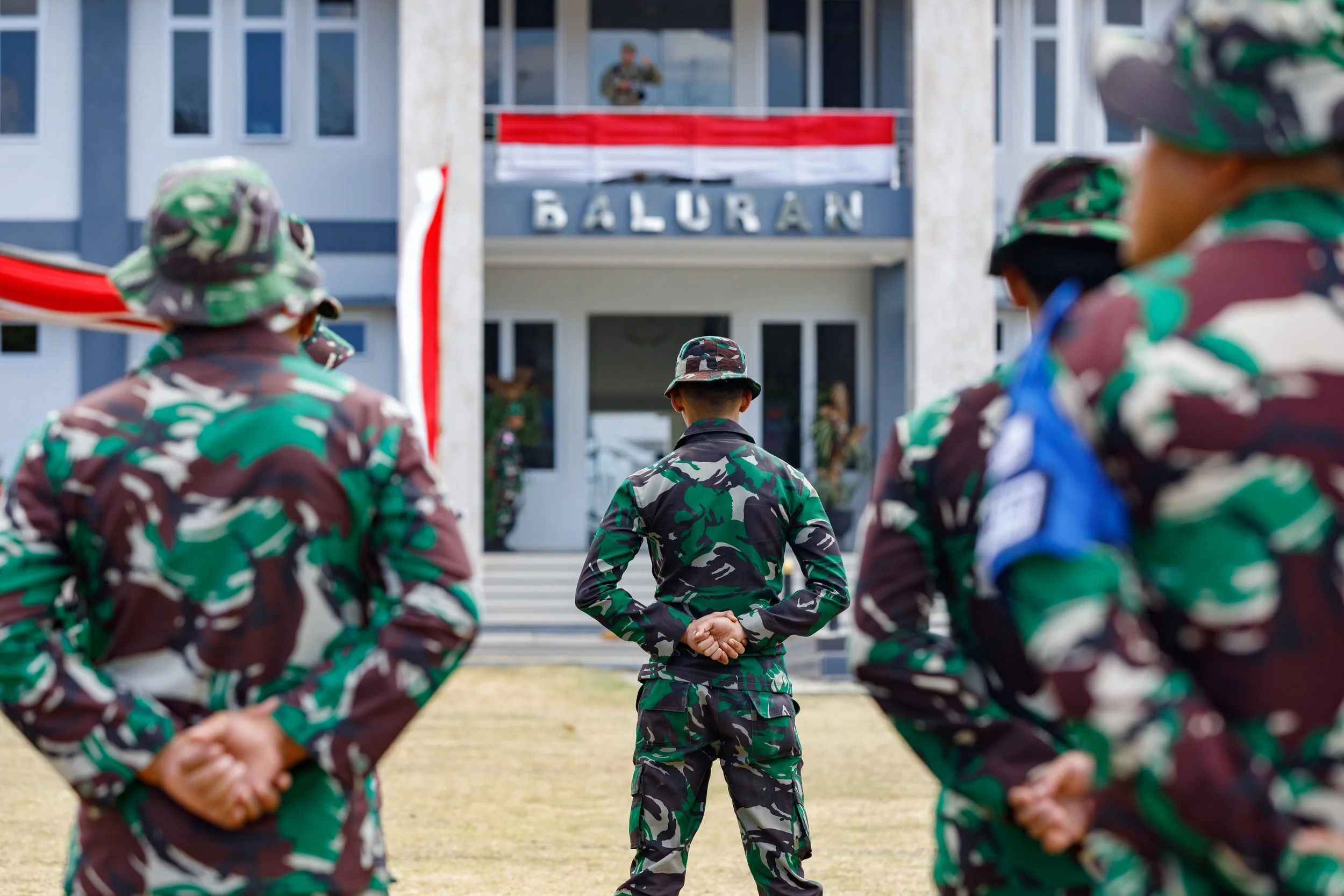 Group of soldiers in camouflage uniforms standing in formation during a ceremony in front of a building with a banner and a soldier speaking at a podium.