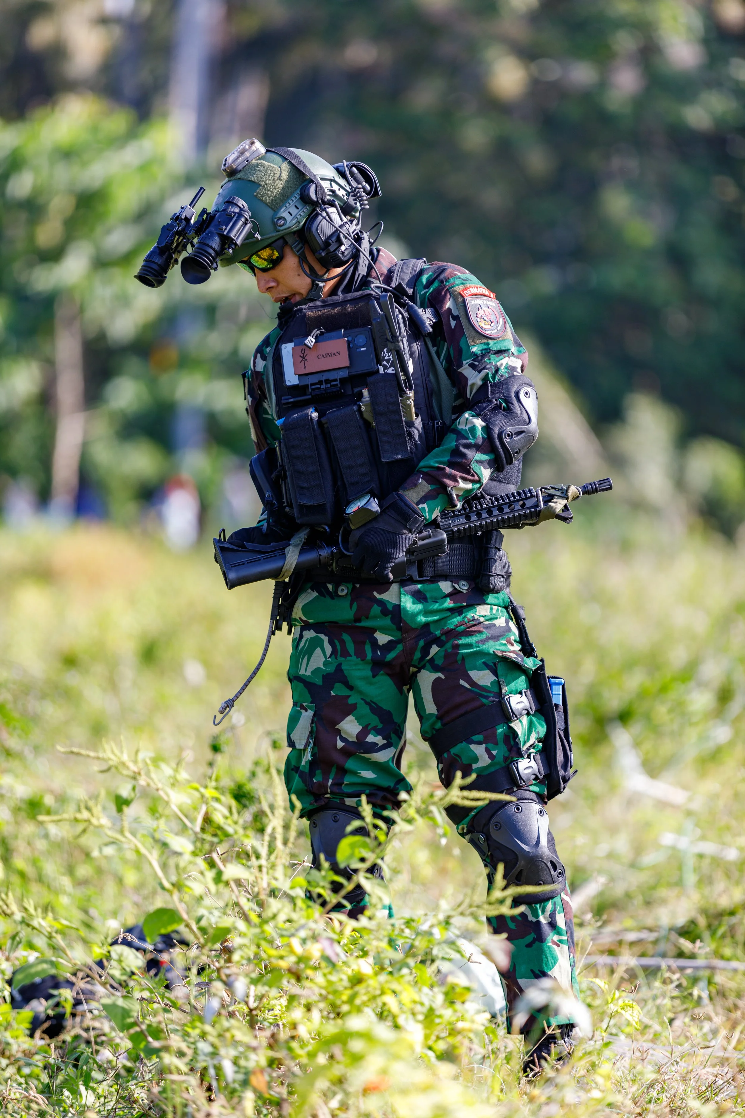 A soldier in camouflage uniform with tactical gear, including a helmet with night-vision goggles, standing in a grassy outdoor area.