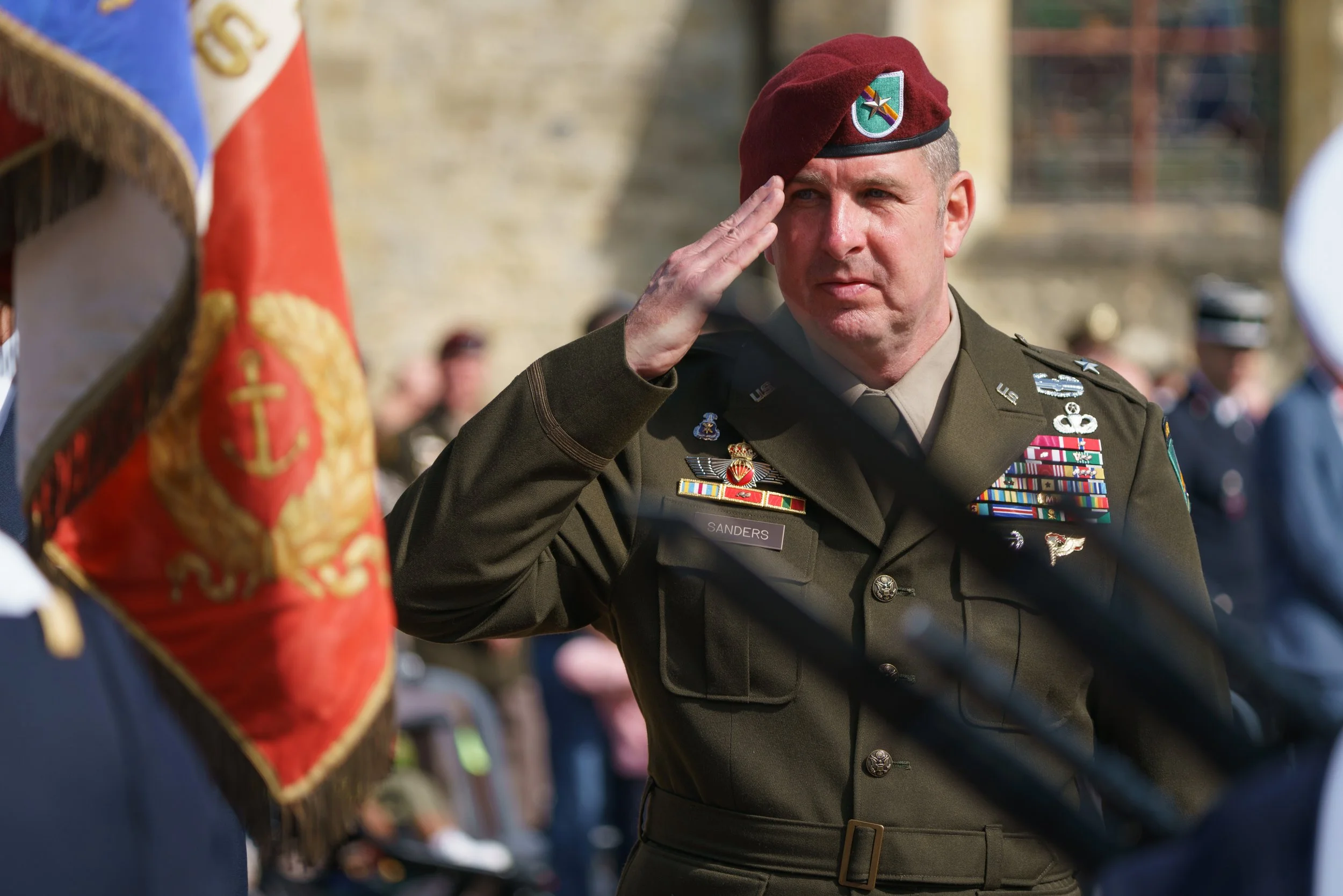 A military officer in uniform salutes during a ceremony, with military decorations on his chest and a red beret, in an outdoor setting with other uniformed personnel and spectators in the background.