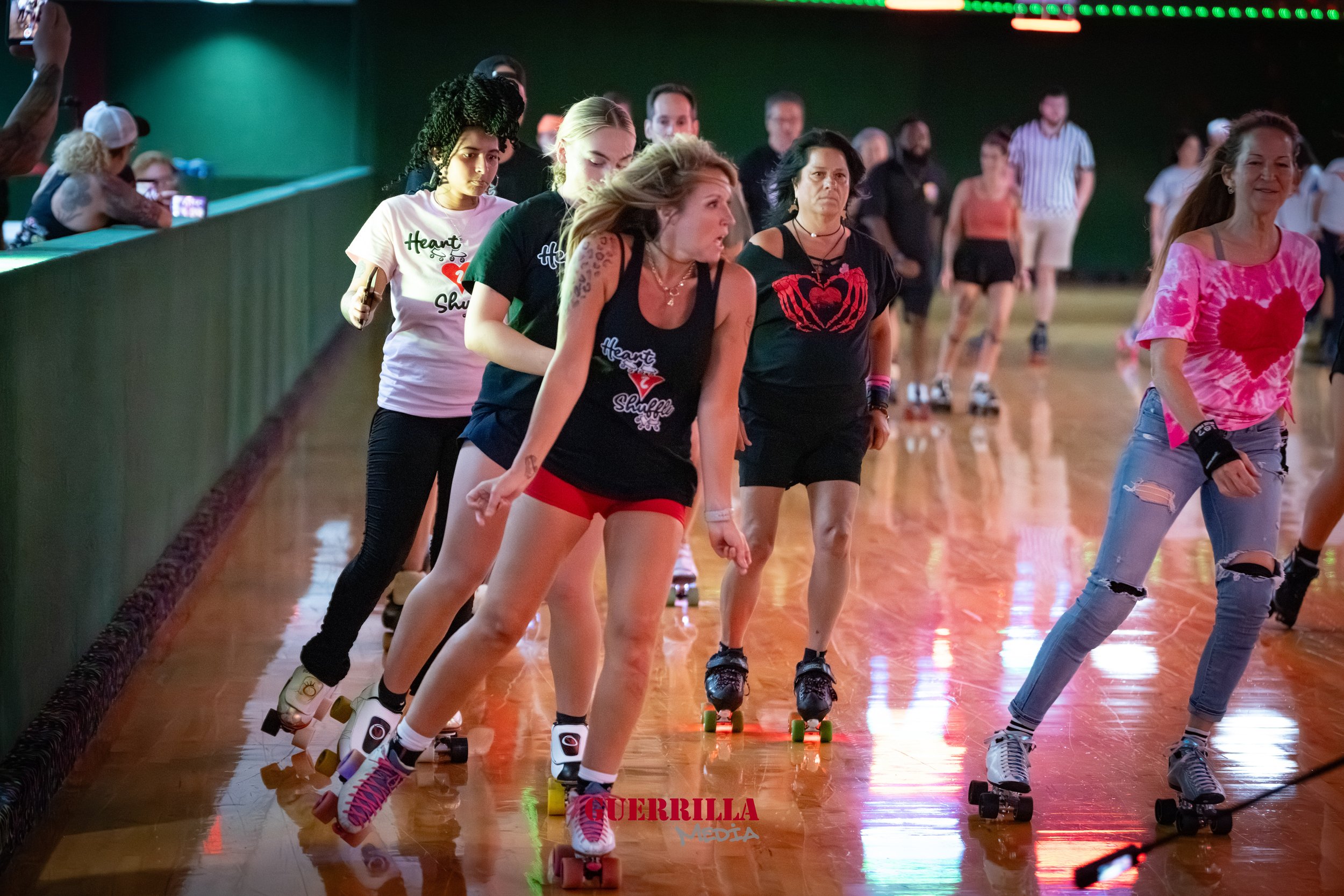 Group of women roller skating indoors, wearing casual athletic clothing, with some wearing team shirts, and a spectator taking photos on the side.