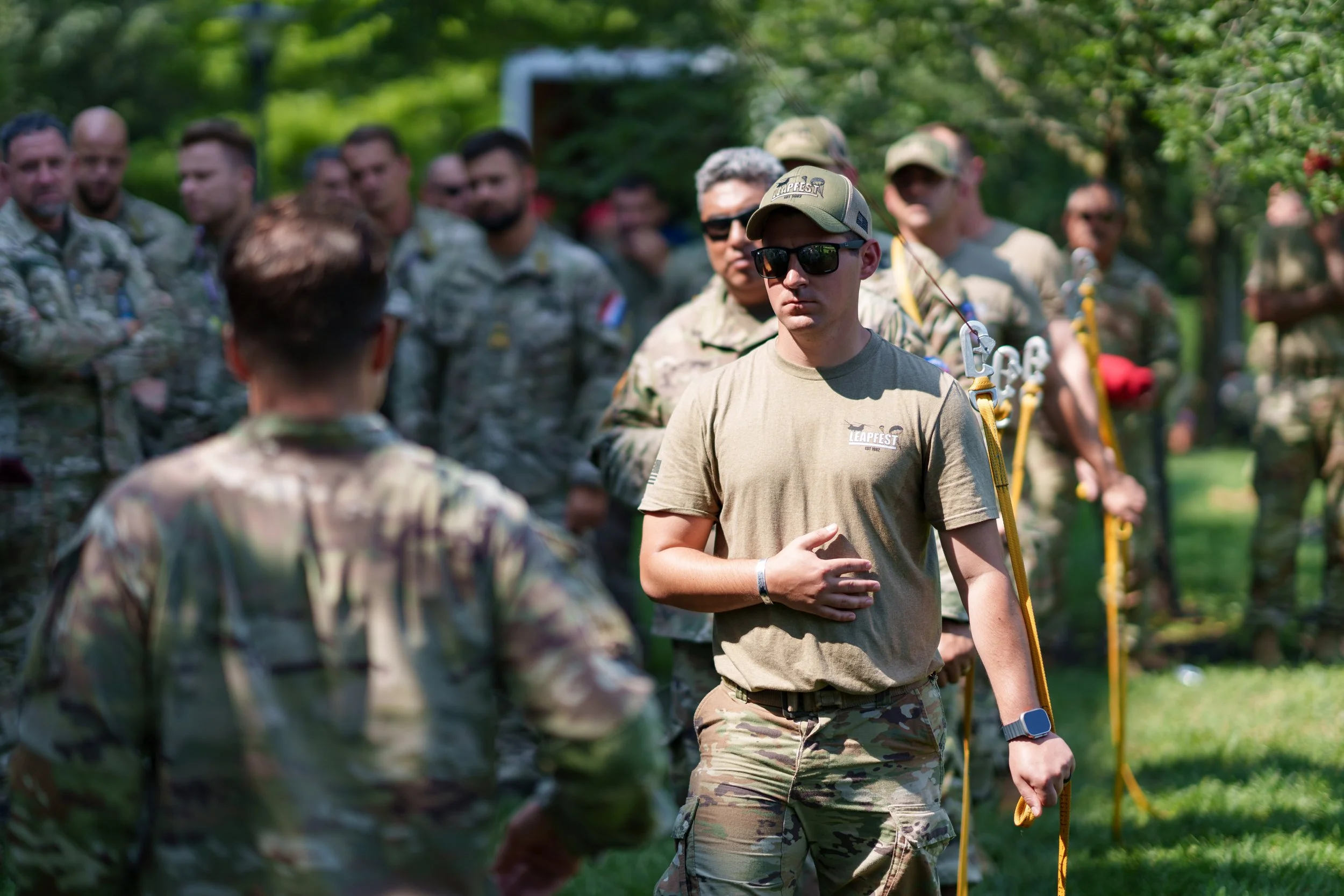 A group of soldiers in camouflage uniforms outdoors, with one soldier in the foreground wearing sunglasses, a tan shirt, and camouflage pants, standing with one hand on his stomach and the other holding a yellow rope.