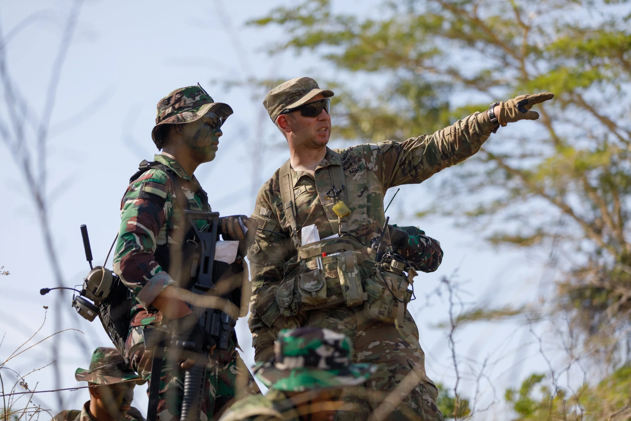 Two military personnel, one in camouflage uniform and the other in tan camouflage uniform, standing outdoors among trees, with one pointing into the distance.