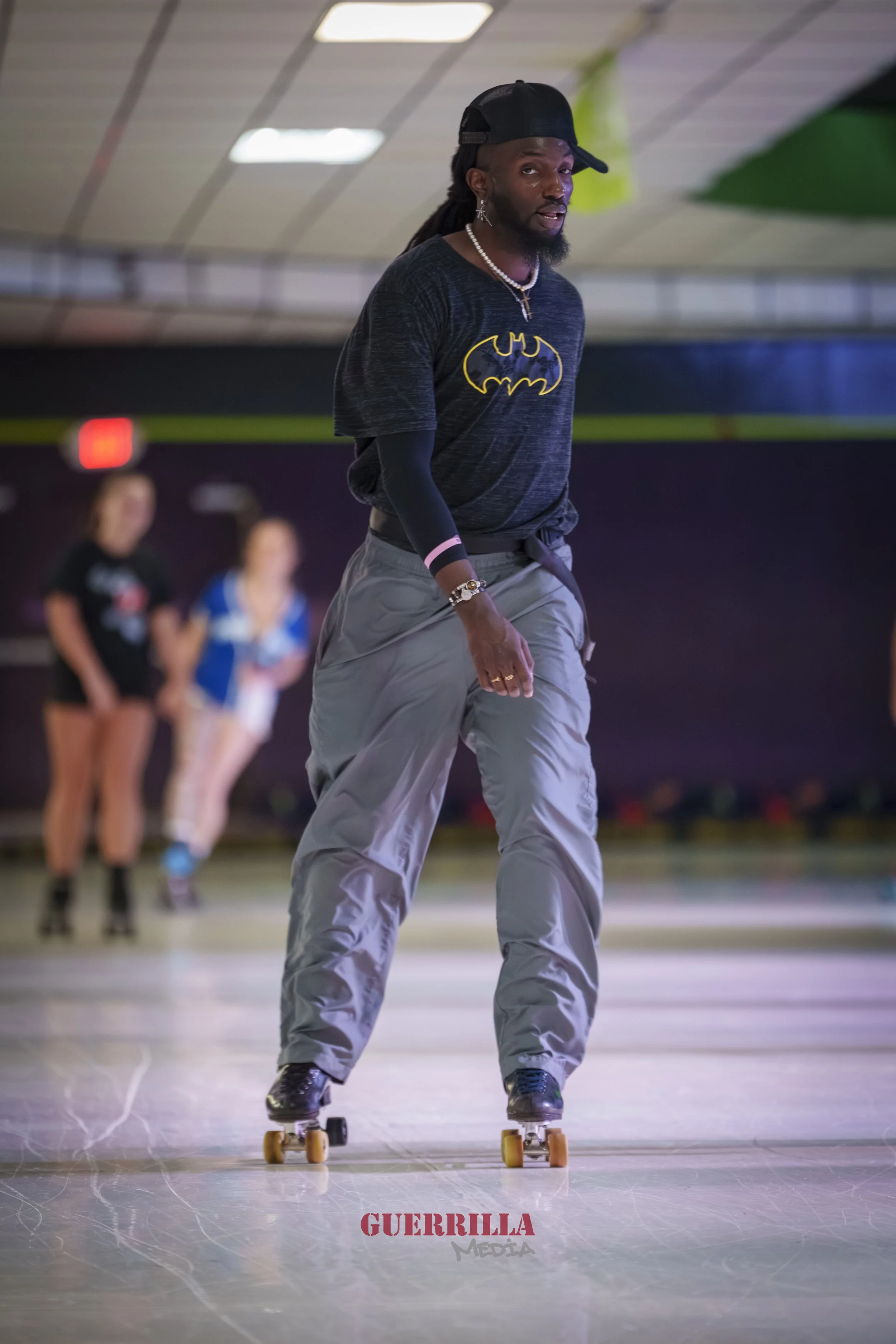 A man roller skating inside a roller rink, wearing a black Batman T-shirt and a black cap, with two women roller skating in the background.