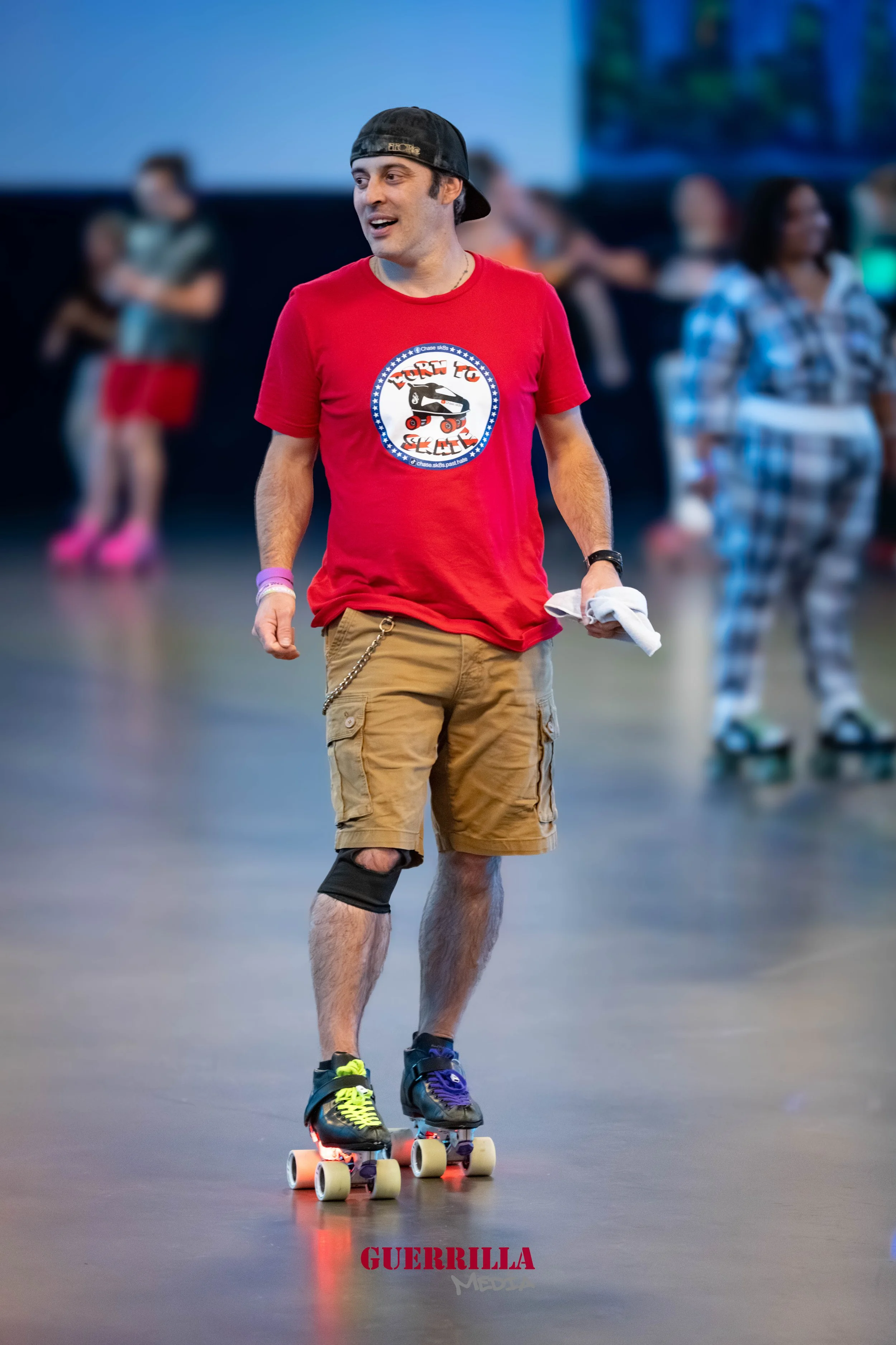 A young man roller skating indoors, wearing a black cap backwards, a red T-shirt with a skate design, tan cargo shorts, and neon-colored skate shoes, holding a cloth, with other people in the background.