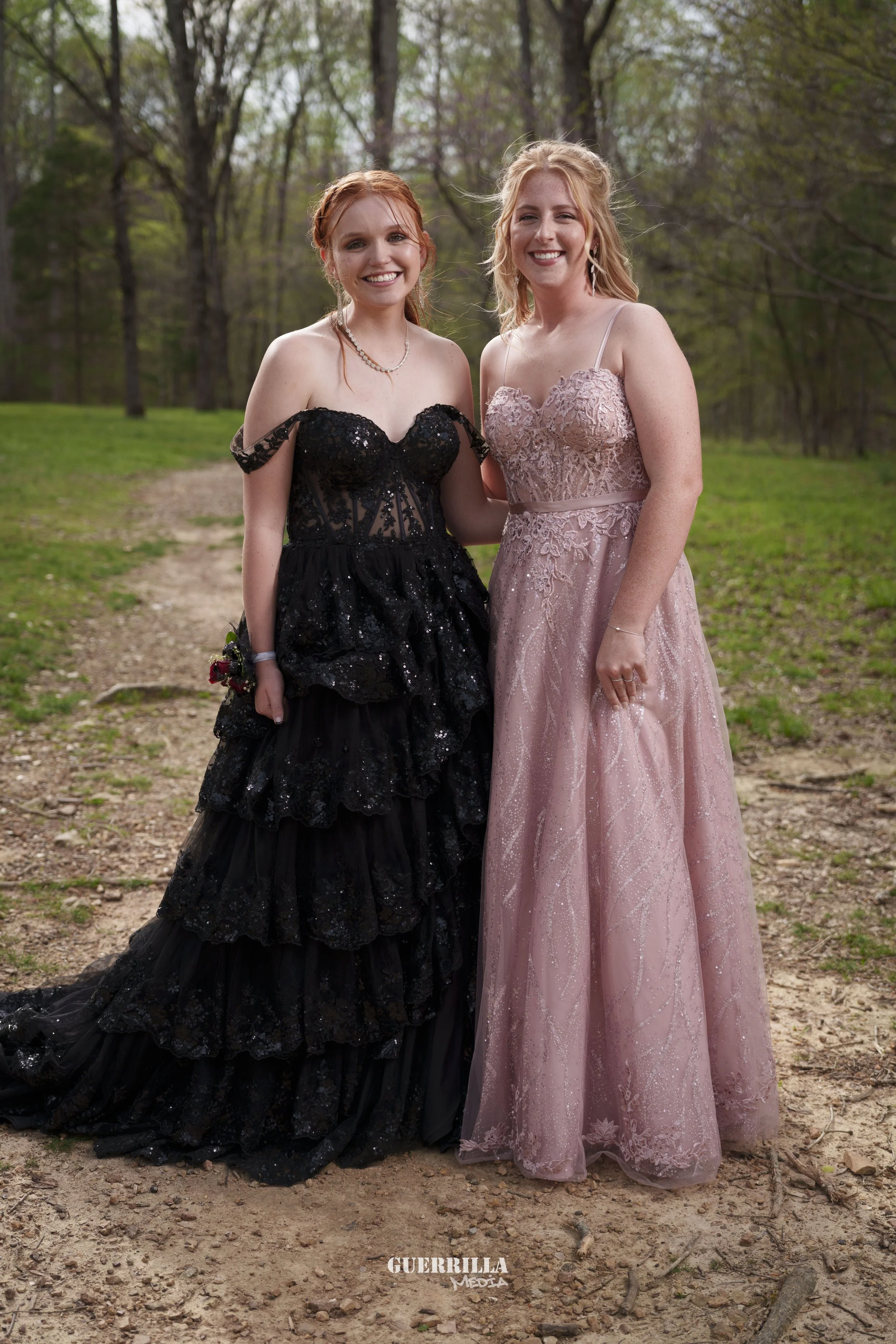 Two young women in formal dresses standing outdoors on a dirt path in a wooded area, smiling at the camera.