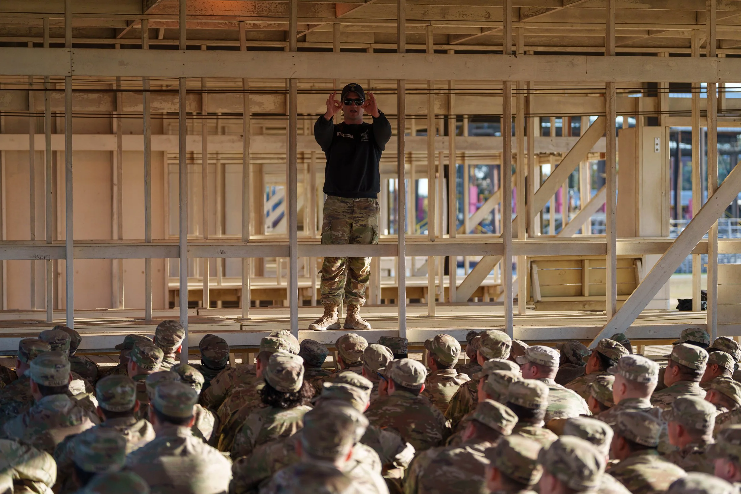 Military instructor speaking to a large group of soldiers seated outdoors during construction of a wooden building.