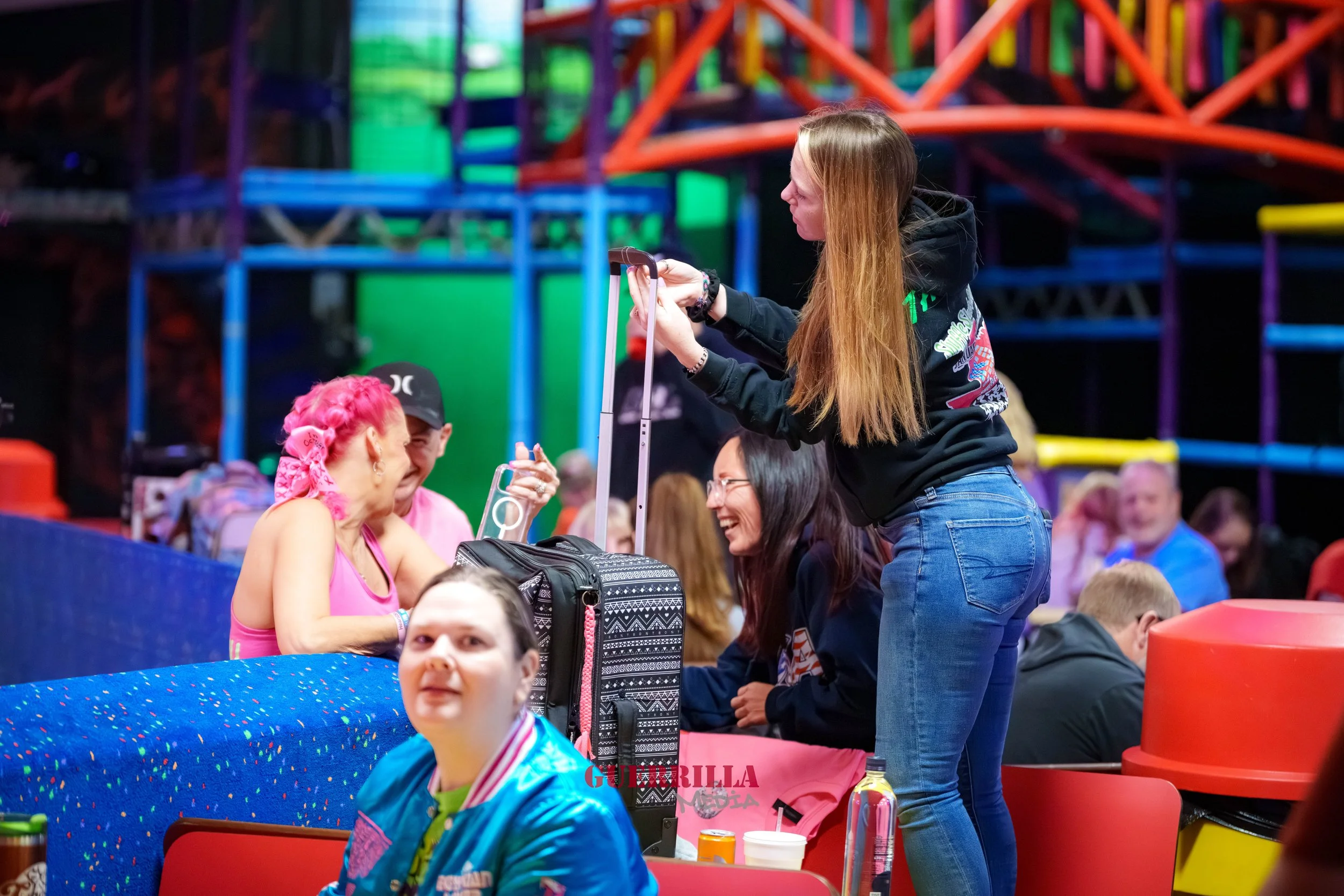 A group of people, including a woman with red hair and pink bandana talking with a woman standing with a suitcase, at an indoor amusement park.