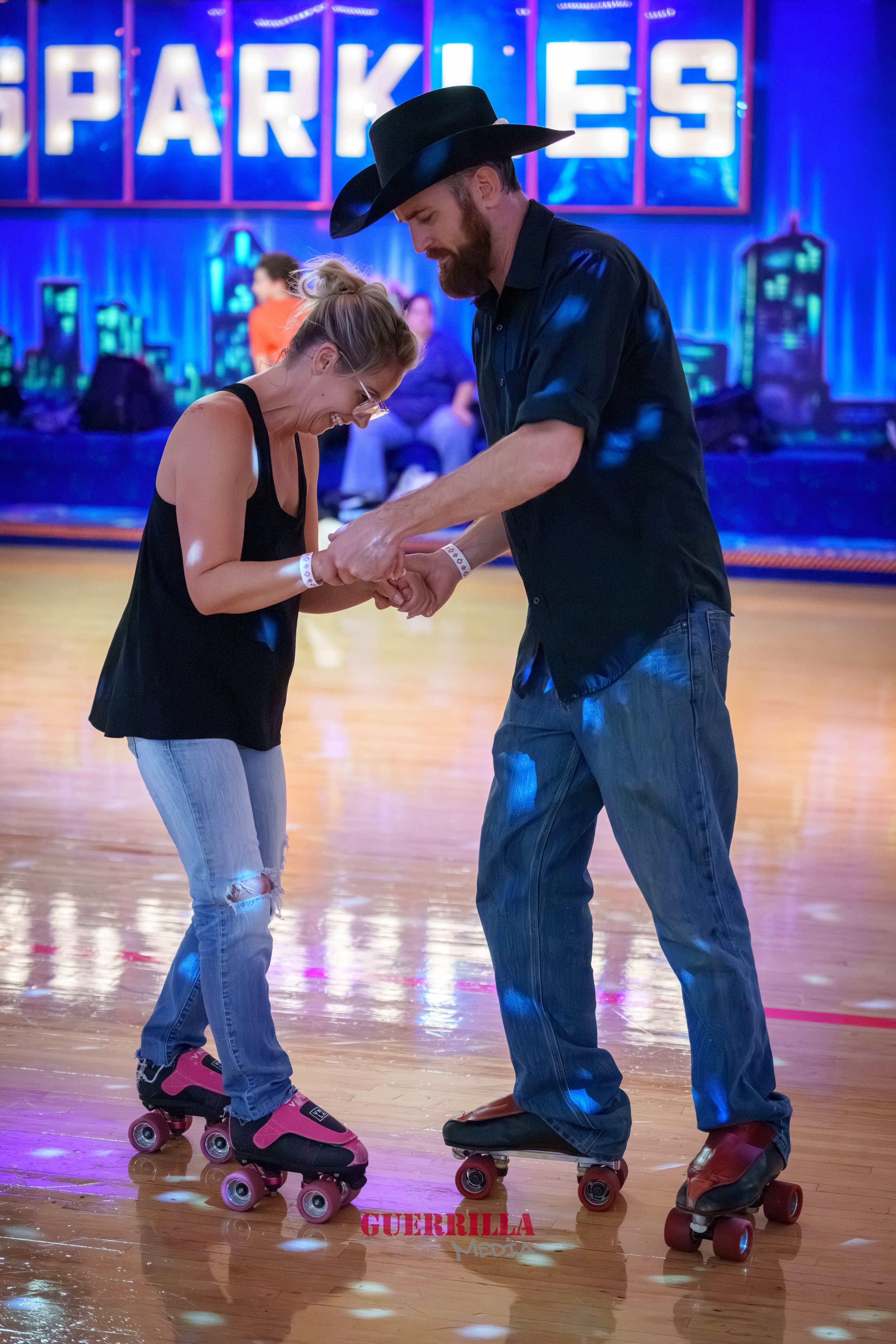 A man and woman roller skating together indoors, with the man holding the woman's hands and both smiling. The woman is wearing a black tank top, ripped jeans, and pink roller skates, while the man is wearing a black shirt, jeans, and cowboy boots. Th