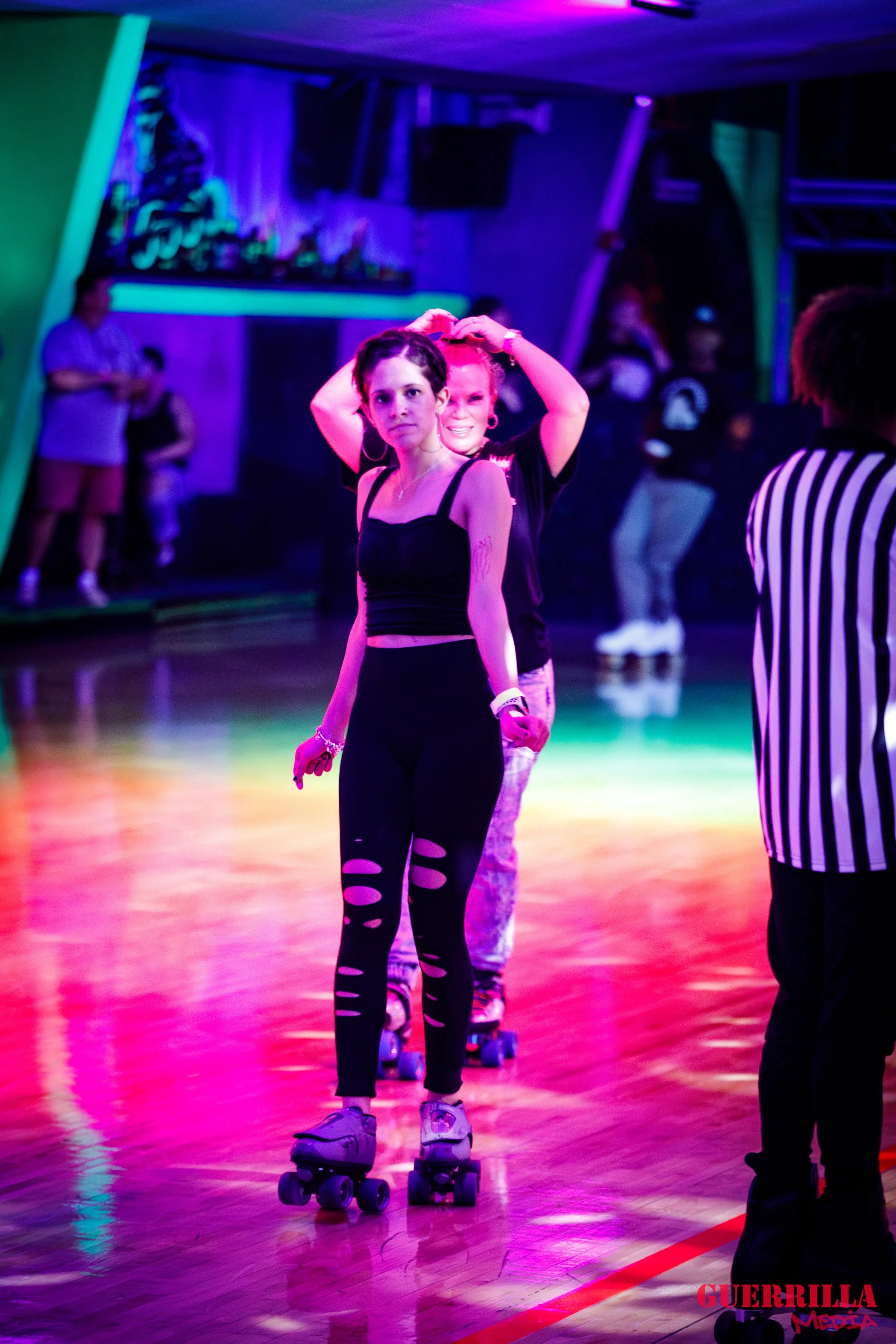 Two women roller skating in a neon-lit indoor skating rink, with other people in the background, one smiling and adjusting her hair.