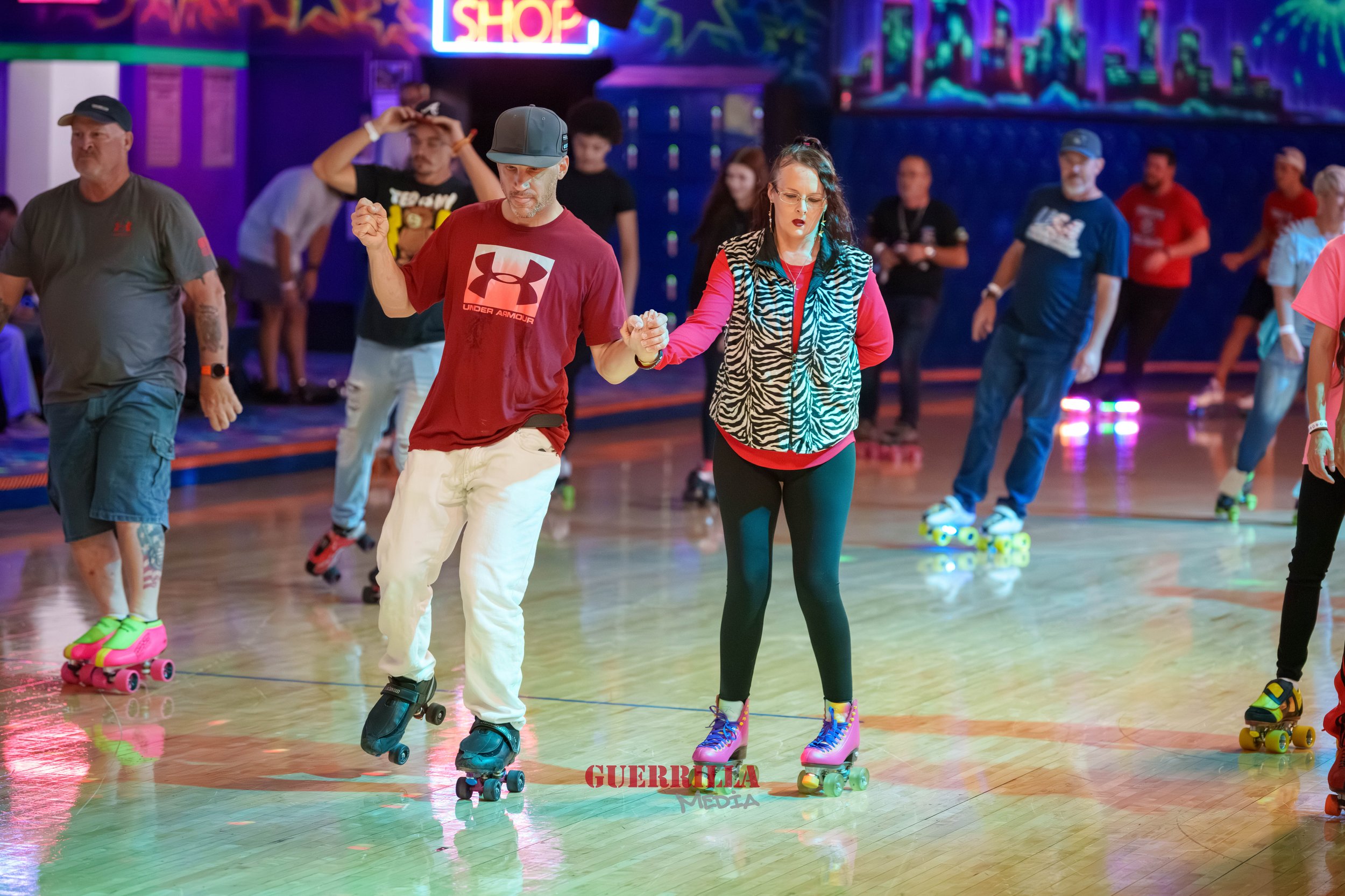 People roller skating indoors in a brightly lit entertainment venue with colorful neon lights, including a woman and man holding hands in the center, surrounded by others skating in the background.