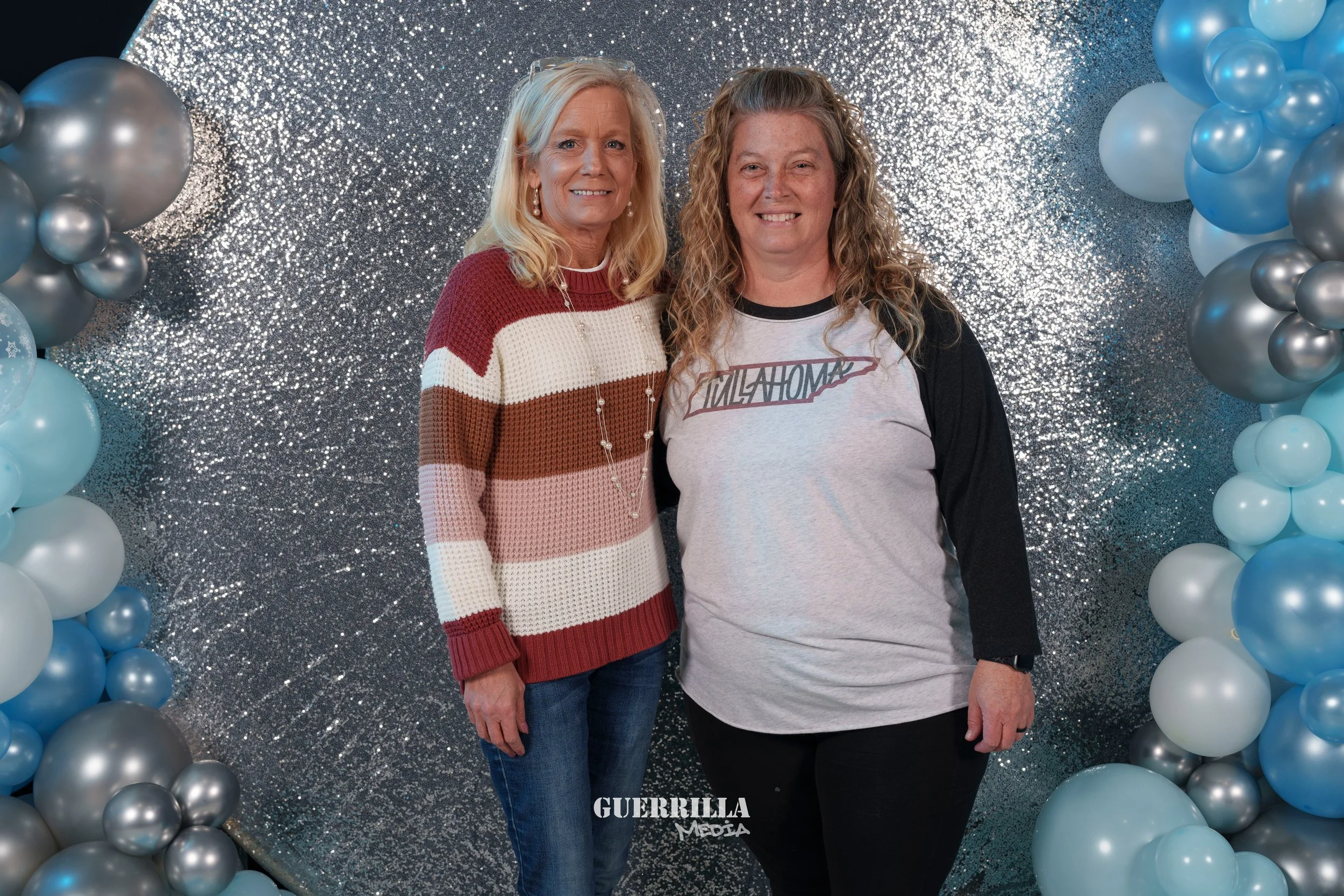 Two women standing side by side in front of a sparkly backdrop with balloon arches on either side. One woman wears a red, white, and brown striped sweater and jeans, while the other wears a gray and black baseball tee with the word 'WILHOM' on it.