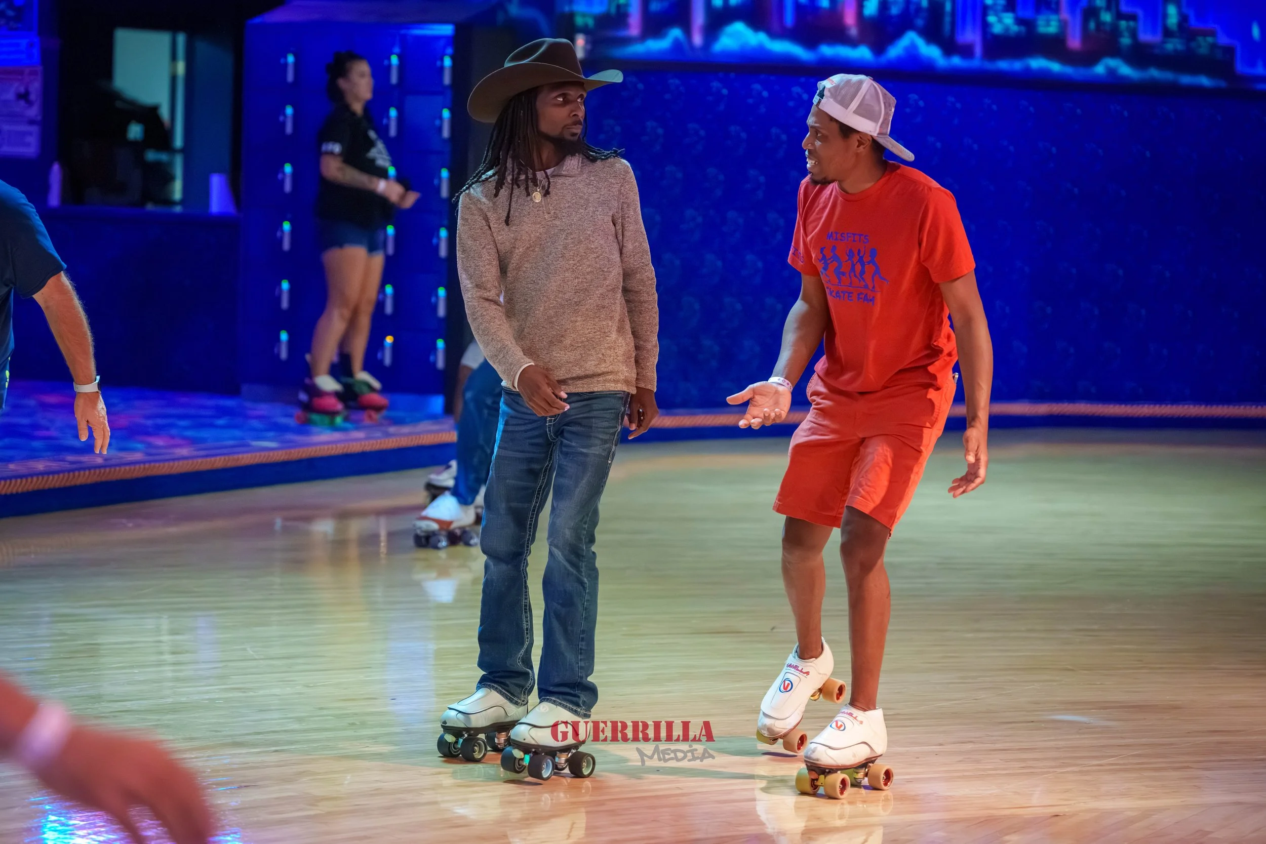 Two men roller skating indoors, one wearing a cowboy hat and beige sweater, the other in a red shirt and cap, engaged in conversation. Background includes other skaters and a blue-lit environment.