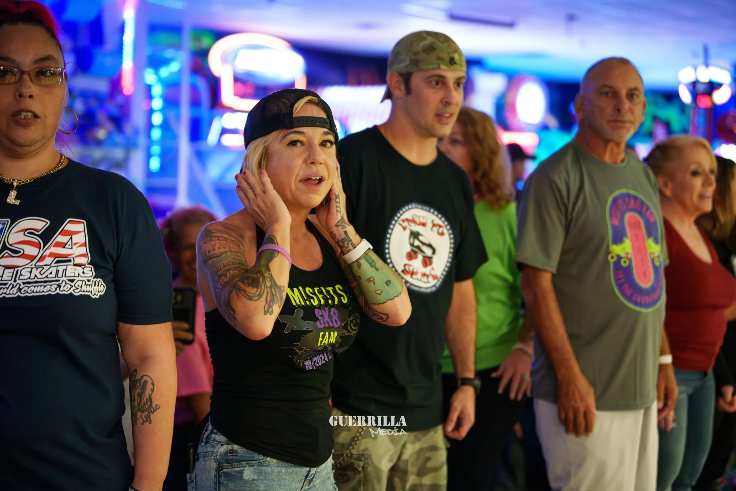 A group of people standing together in a colorful indoor setting, some wearing band or event t-shirts, and a woman with tattoos covering her arms, wearing a black tank top and a backwards cap, touching her ears and looking surprised or concerned.
