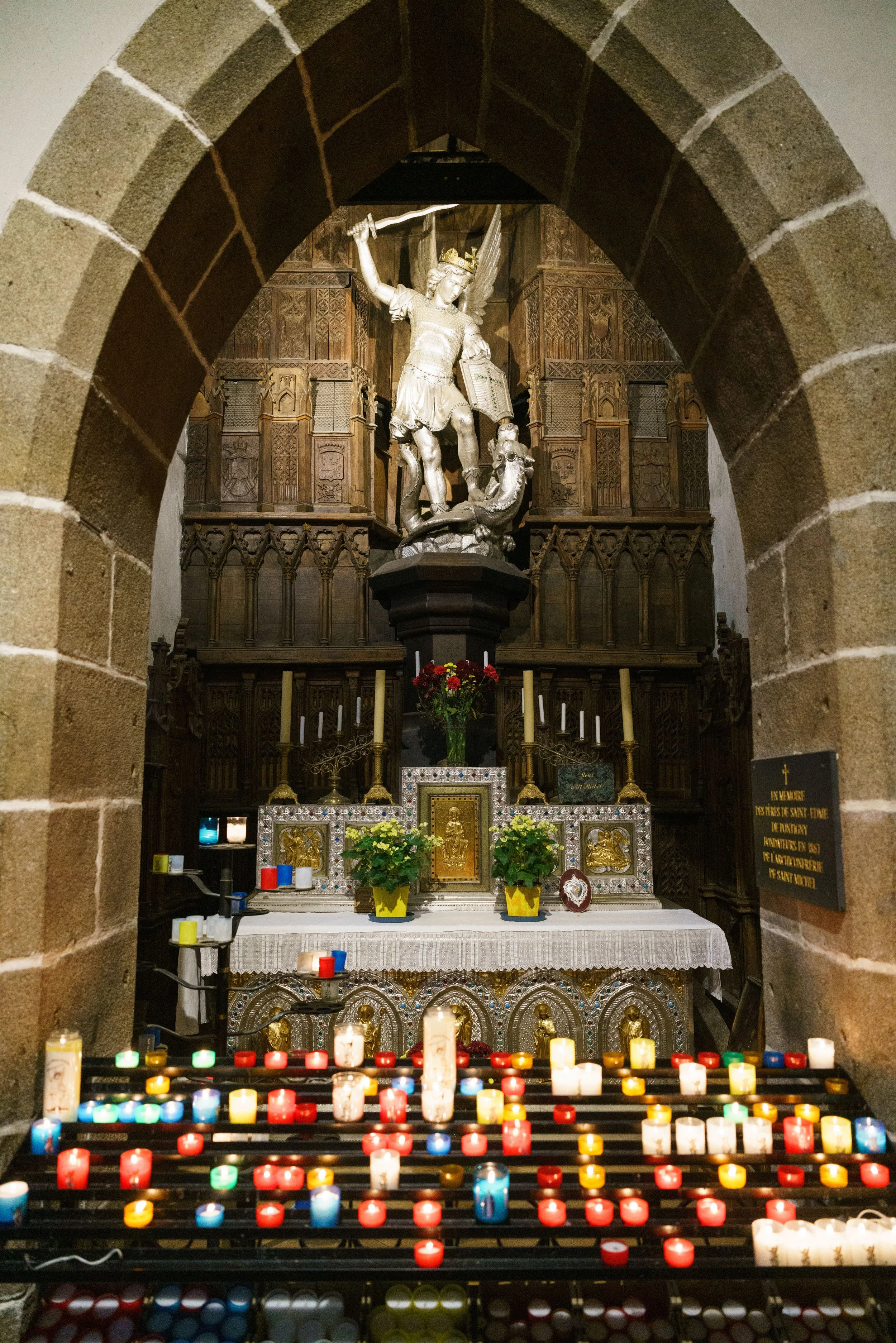 Inside a church, there is a stone archway leading to an altar with a statue of Saint Michael the Archangel slaying a demon, surrounded by candles and flowers.