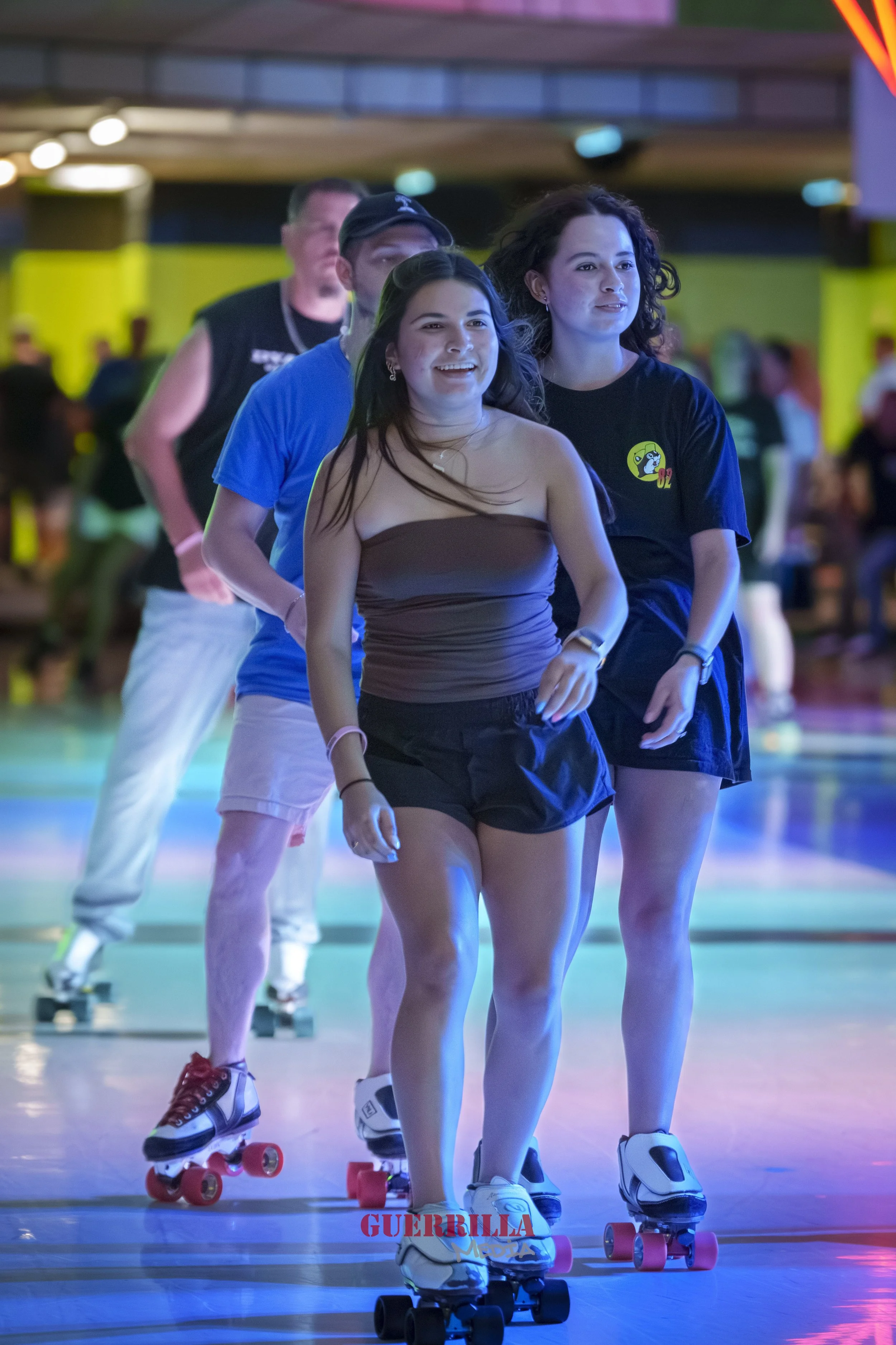 Four people roller skating indoors at a rink. The first girl is smiling, wearing a brown tube top and black shorts. The second girl has curly hair, wearing a black T-shirt and shorts. Two men are in the background, one wearing a blue T-shirt and gray