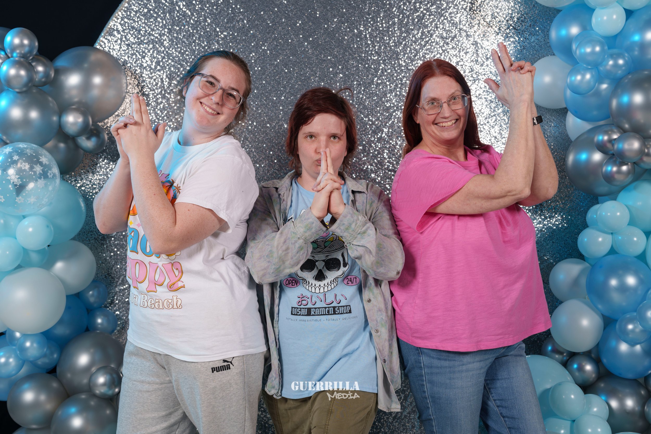 Three women posing with playful expressions in front of a silver glitter backdrop, surrounded by blue and silver balloons.
