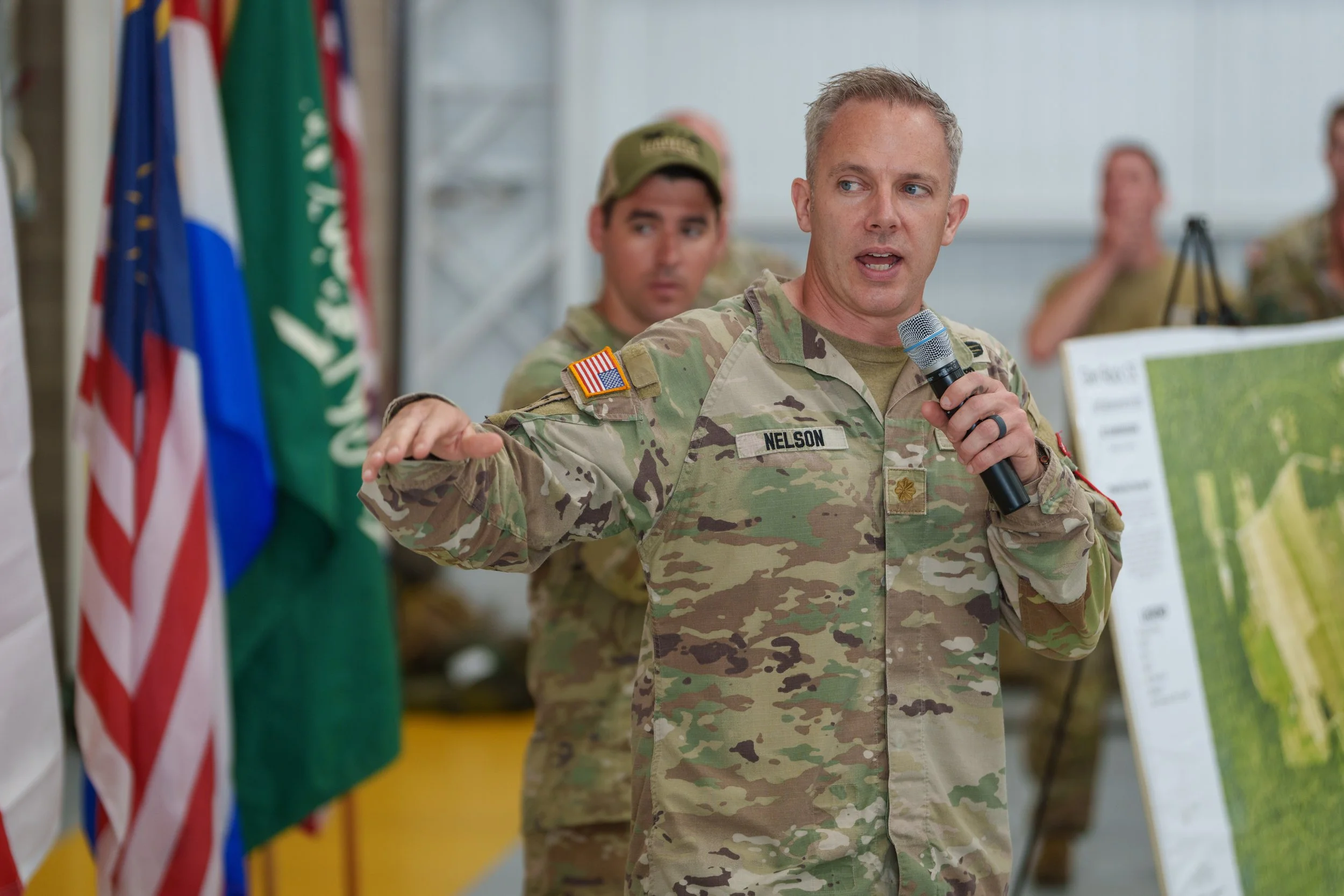 A man in military uniform holding a microphone and speaking, with several other soldiers and flags in the background.
