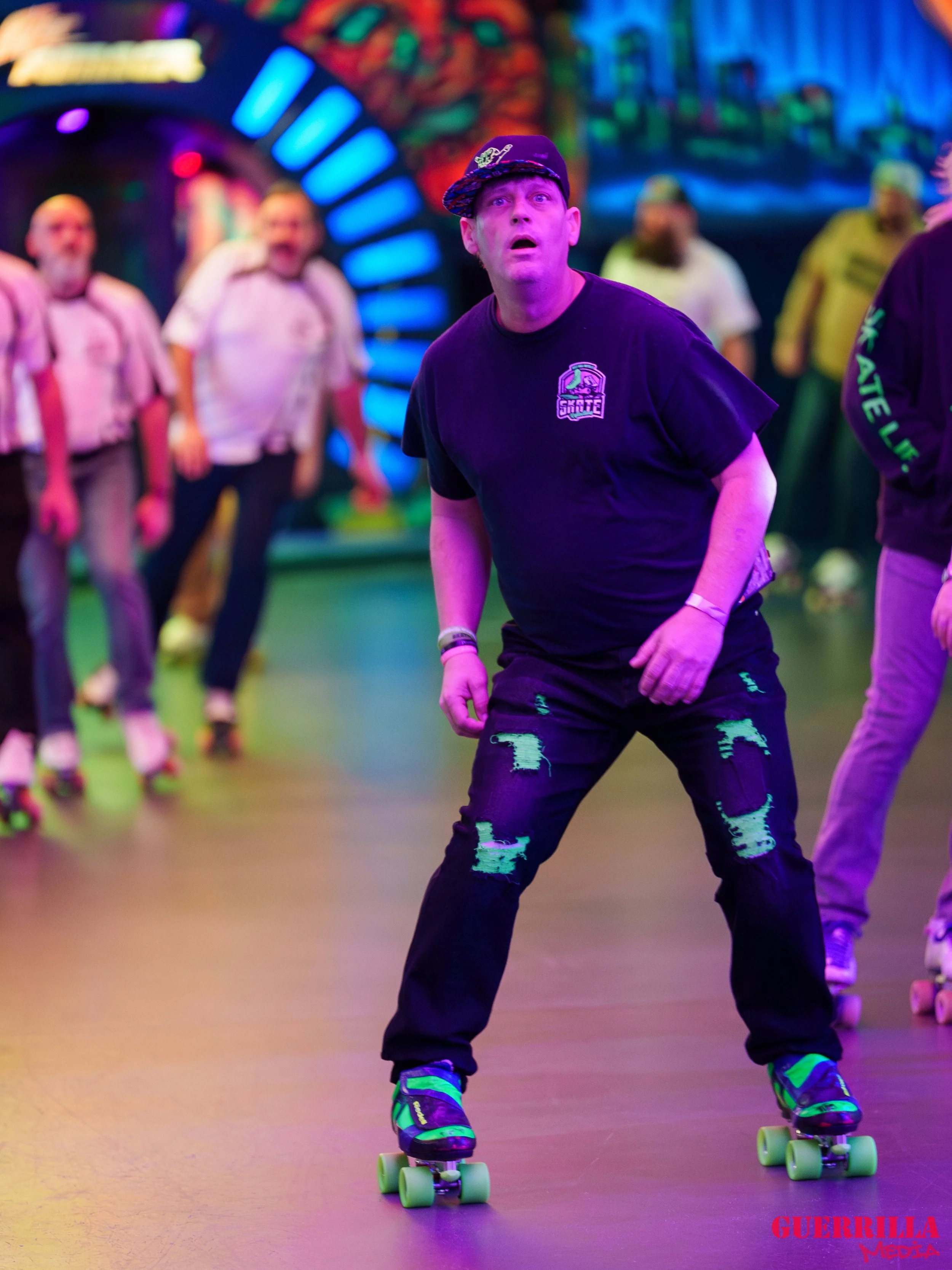 A man with a surprised expression on his face skating inside an amusement park or roller rink, with other skaters and colorful lights in the background.