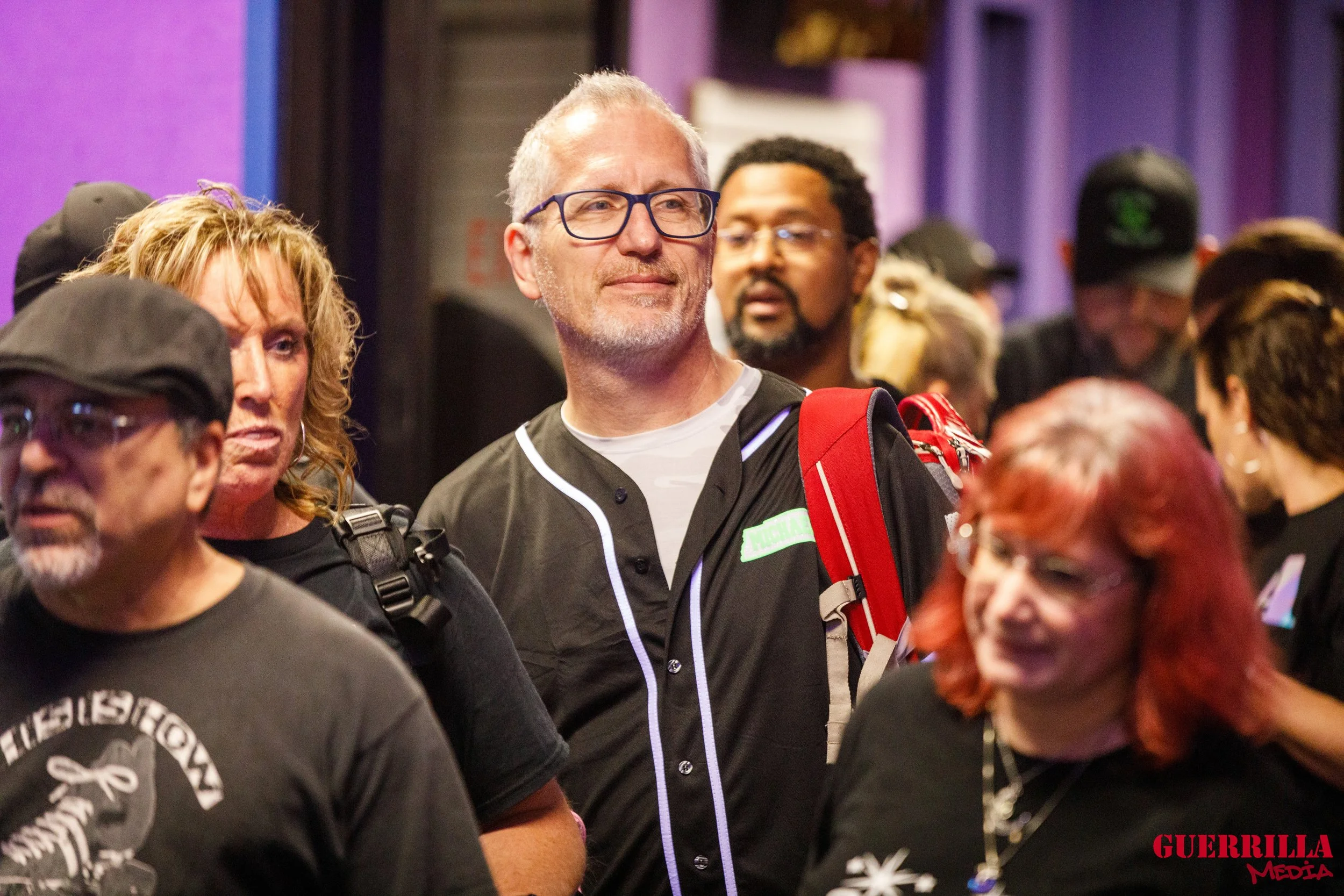 A group of people gathered at an indoor event, with a man wearing glasses and a black jacket at the center, surrounded by others, some with backpacks and casual clothing.