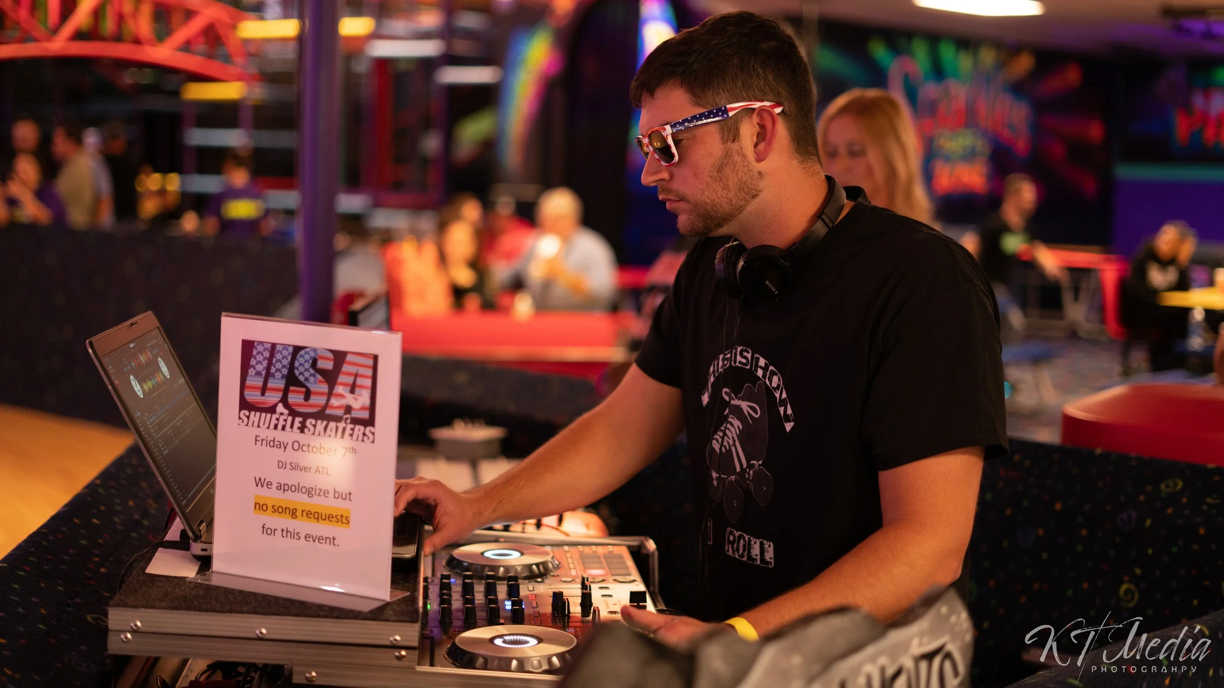 A DJ wearing patriotic sunglasses and headphones is playing music at an event with a sign that reads, "USA Shuffle Skaters, Friday October 7th, DJ Silver ATL, We apologize but no song requests for this event."