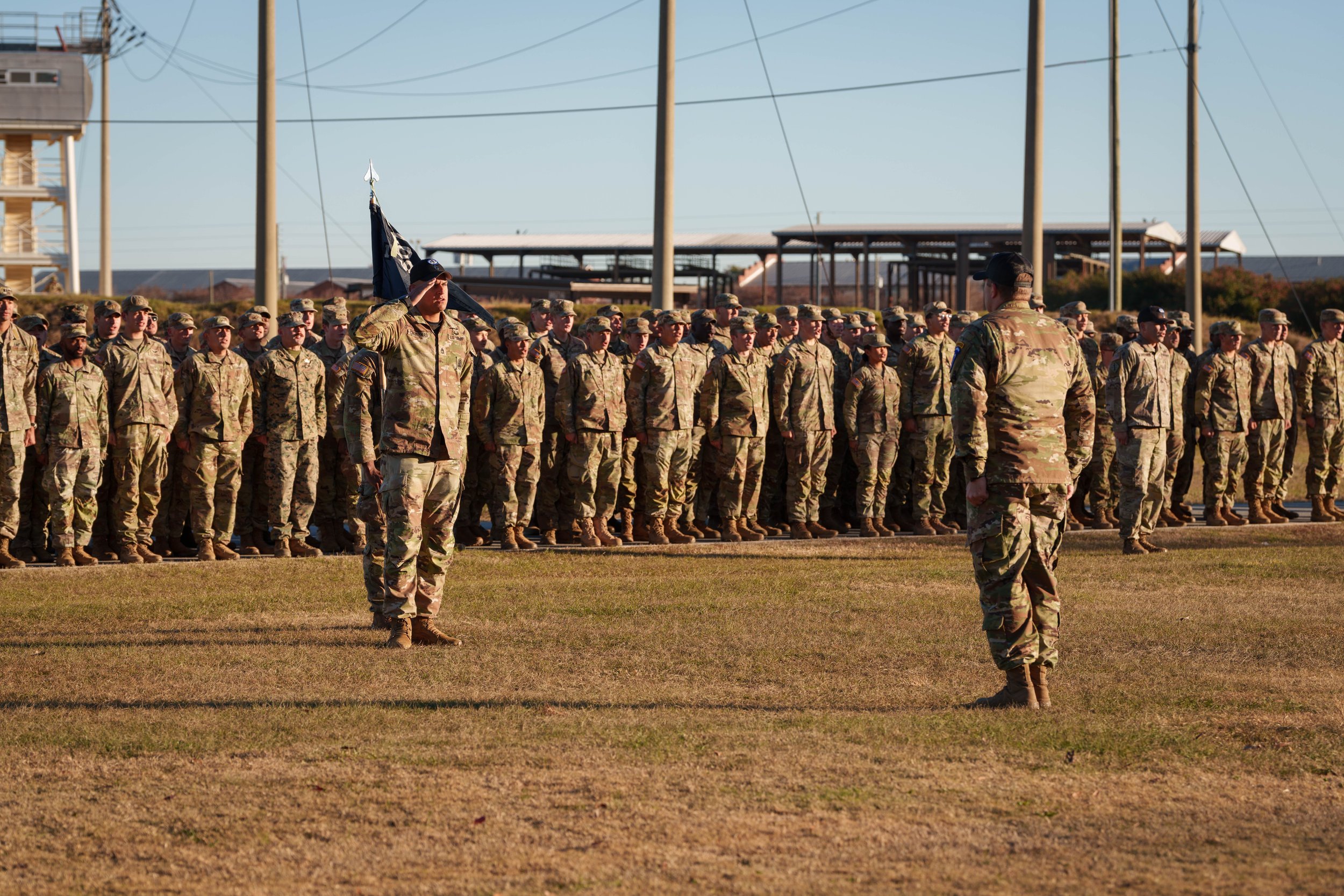 Military formation with soldiers standing in formation on a grassy field, with a commanding officer saluting a soldier holding a flag. Background includes utility poles, buildings, and clear sky.