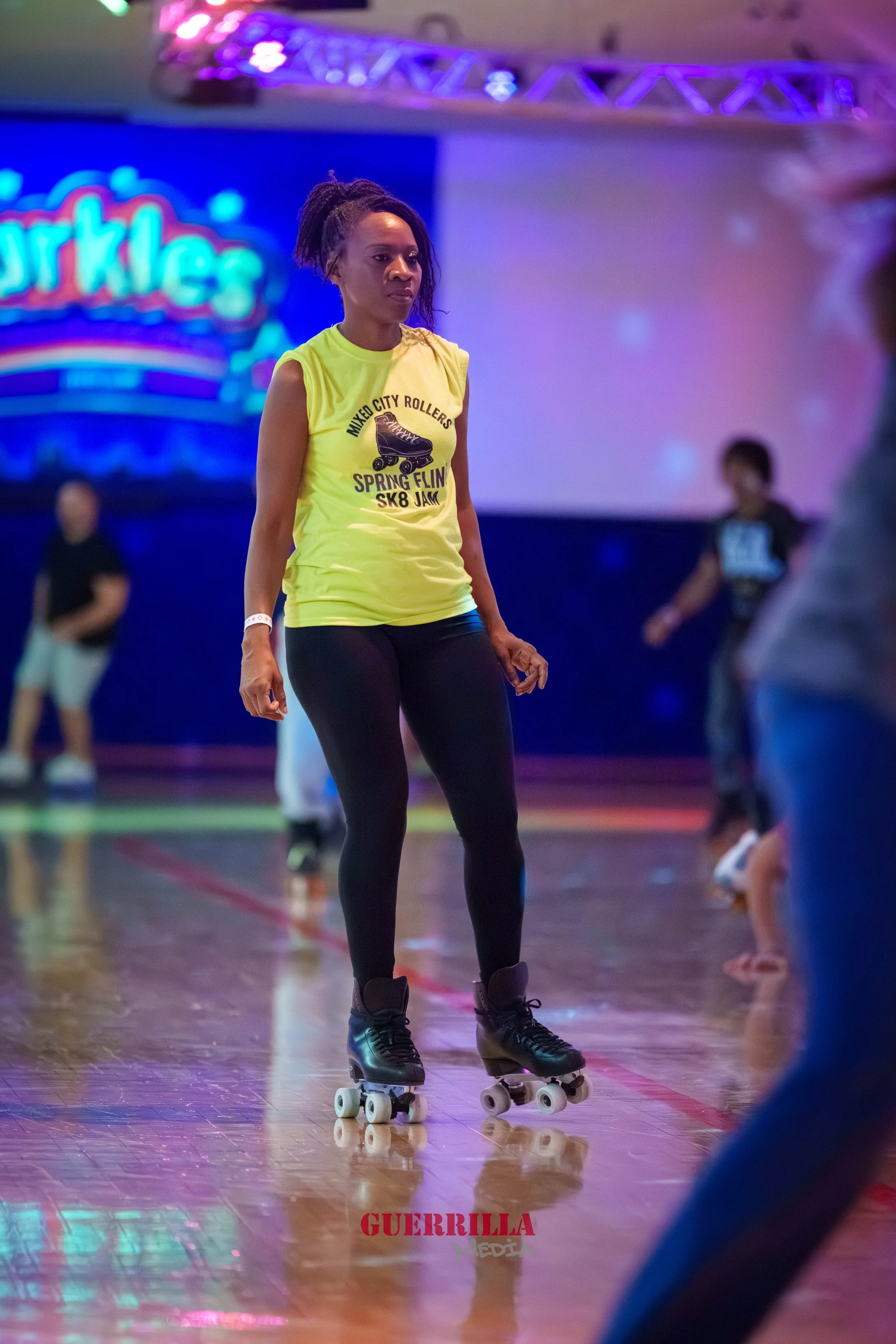 A woman roller skating on a wooden floor at an indoor roller skating rink, wearing a yellow sleeveless shirt and black leggings, with other skaters in the background.