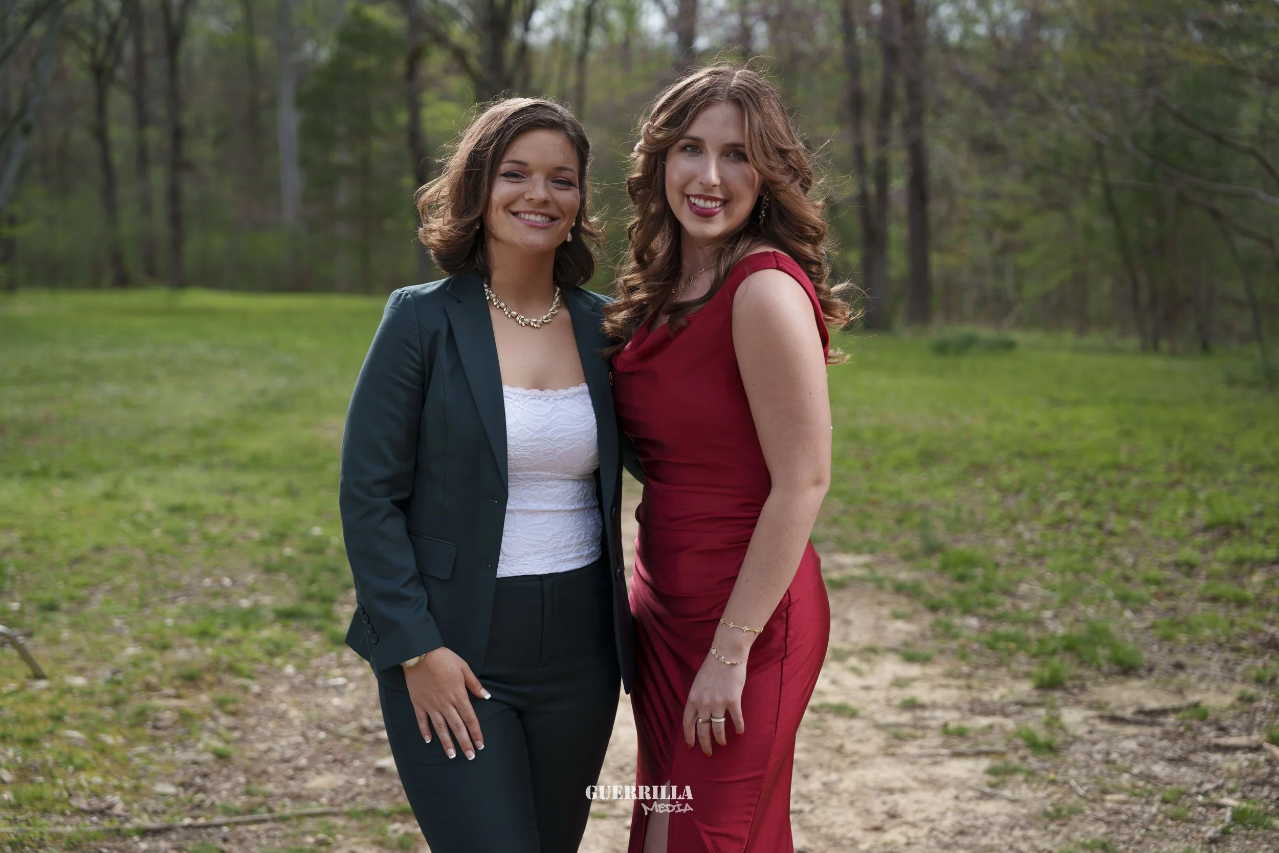 Two women standing outdoors on a dirt path in a wooded area, smiling at the camera, one wearing a dark blazer with a white top, the other in a red dress.