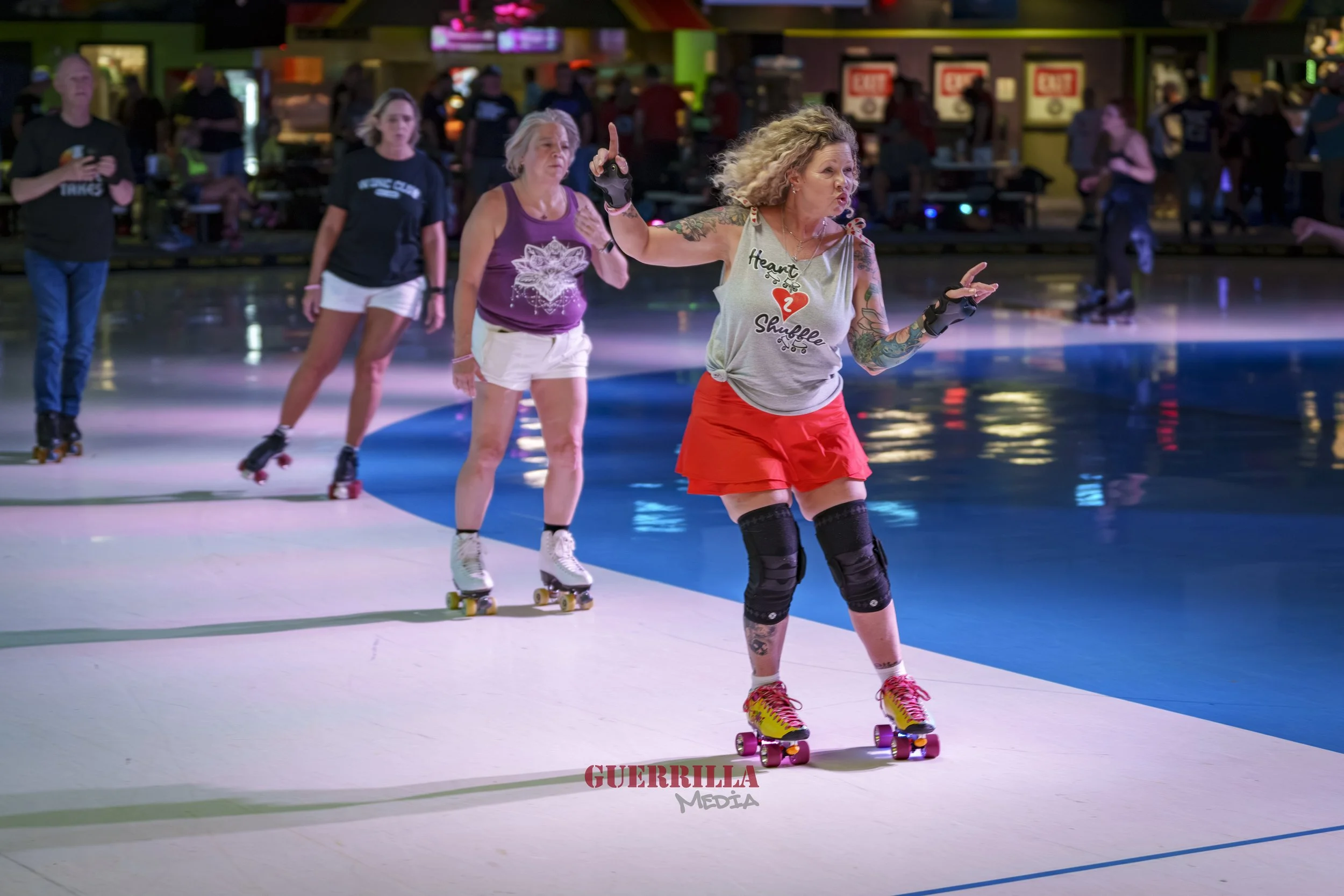 A group of women roller skating at an indoor rink with colorful lighting and people in the background, with one woman in the foreground wearing a gray tank top, red skirt, and knee pads.