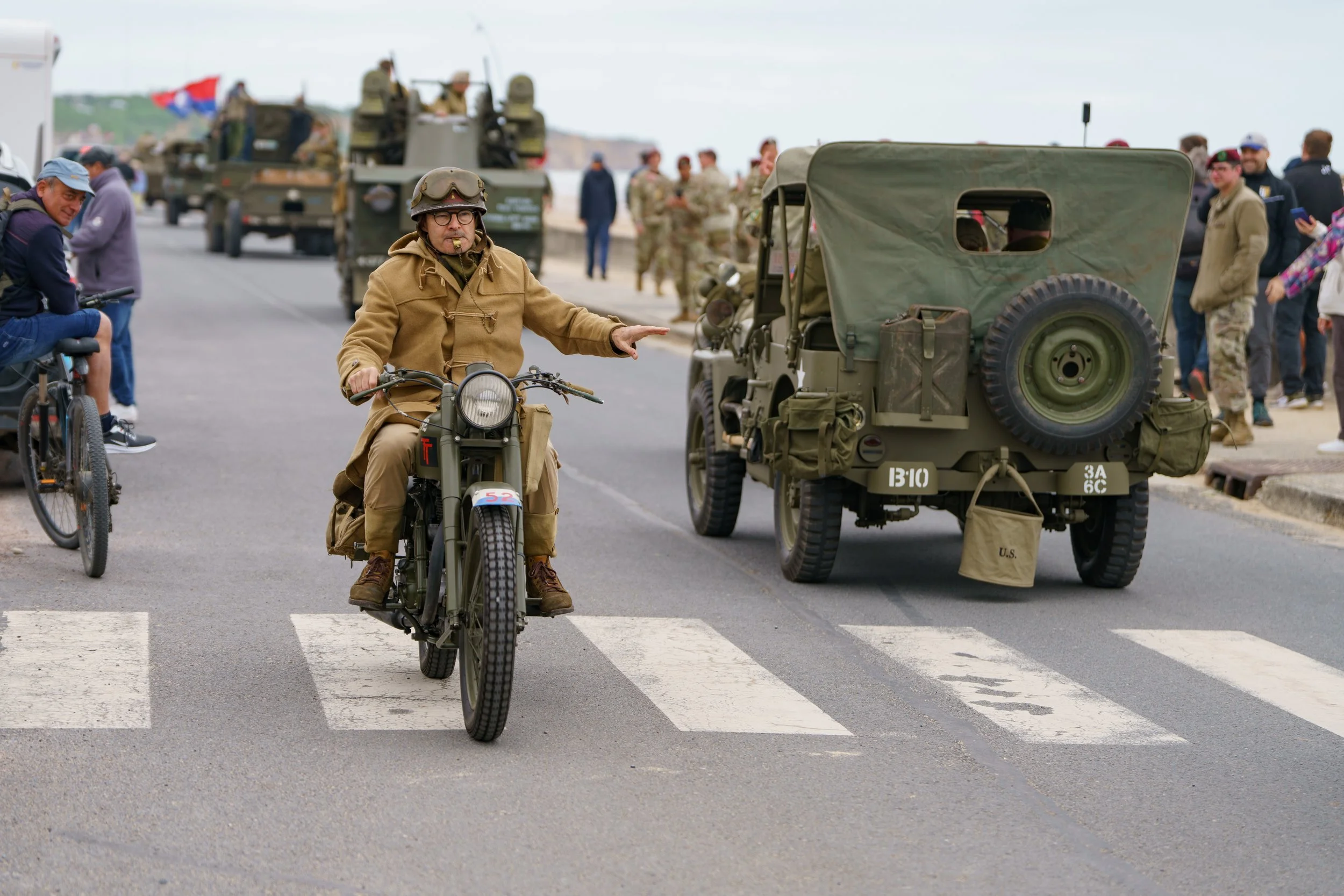 A man riding a motorcycle across a crosswalk during a military event, with a vintage military vehicle and people watching in the background.