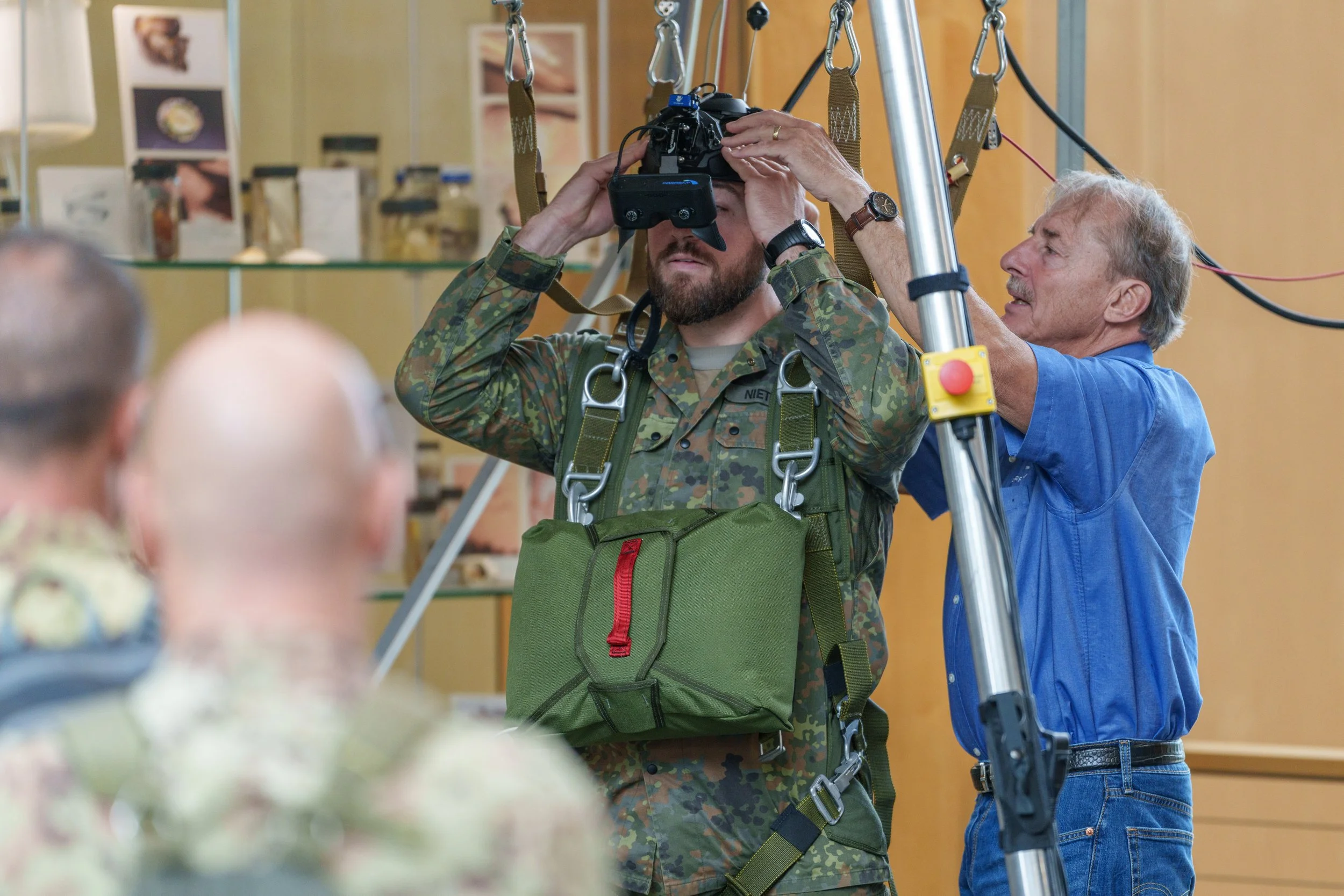 A man in military camouflage gear is wearing a virtual reality headset, assisted by an older man in a blue shirt adjusting equipment on a setup with a metal stand and wires. Blurred people are in the foreground, and shelves with pictures and objects 