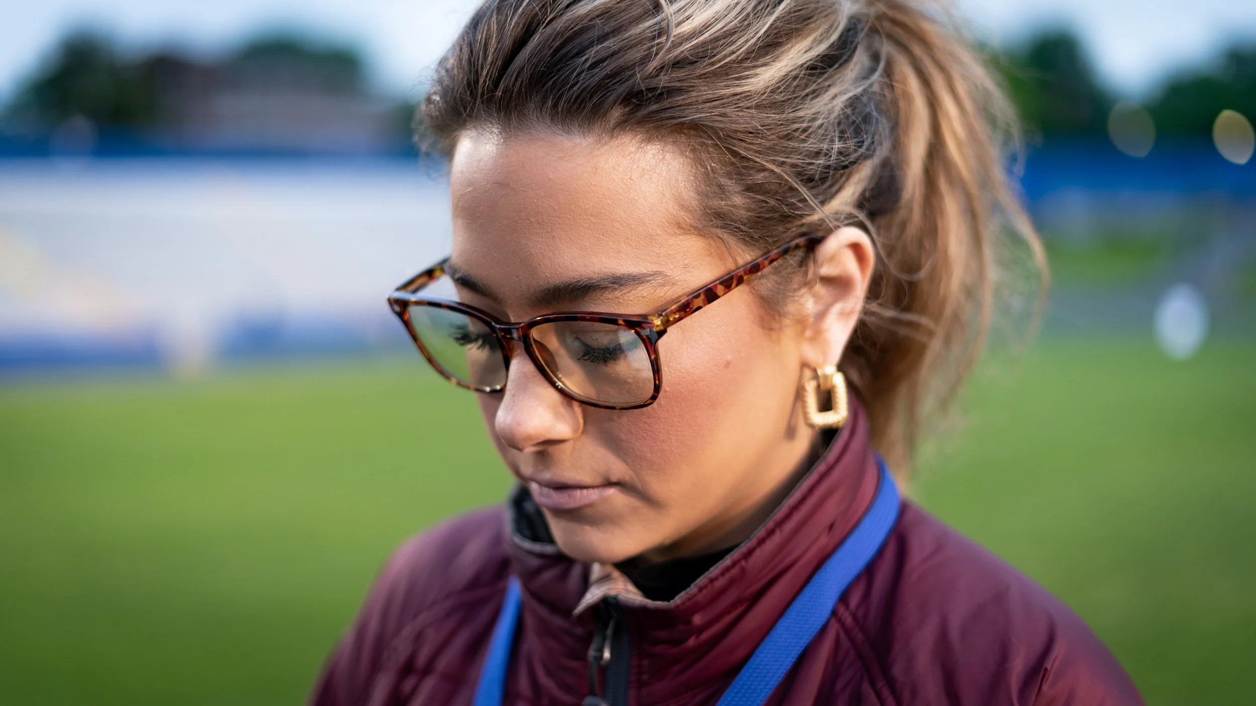 Close-up of a woman with curly brown hair wearing tortoiseshell glasses, gold earrings, and a maroon jacket, with a blurred outdoor background.