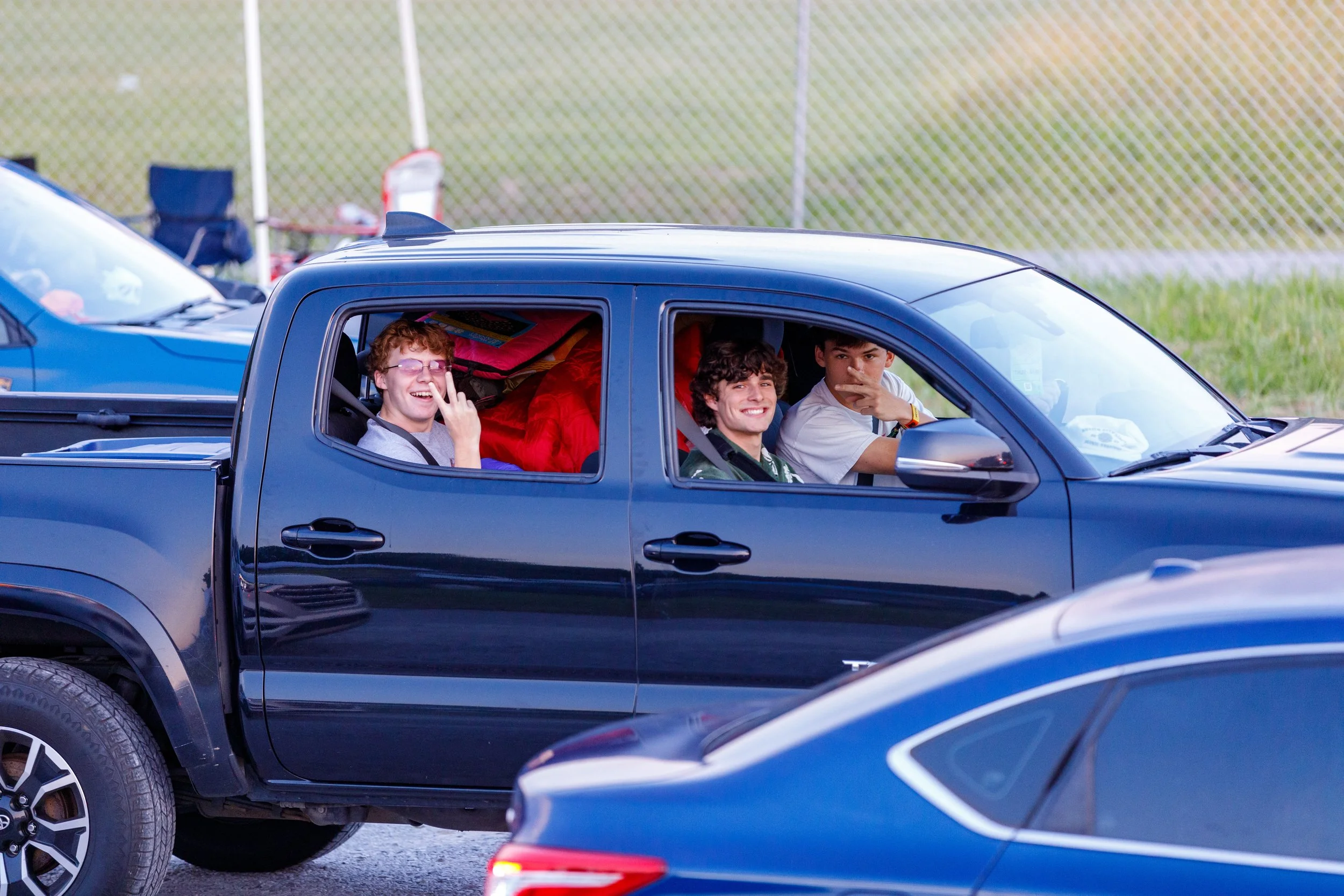 Three young men sitting in a black pickup truck, smiling and making gestures, parked outdoors near a chain-link fence.