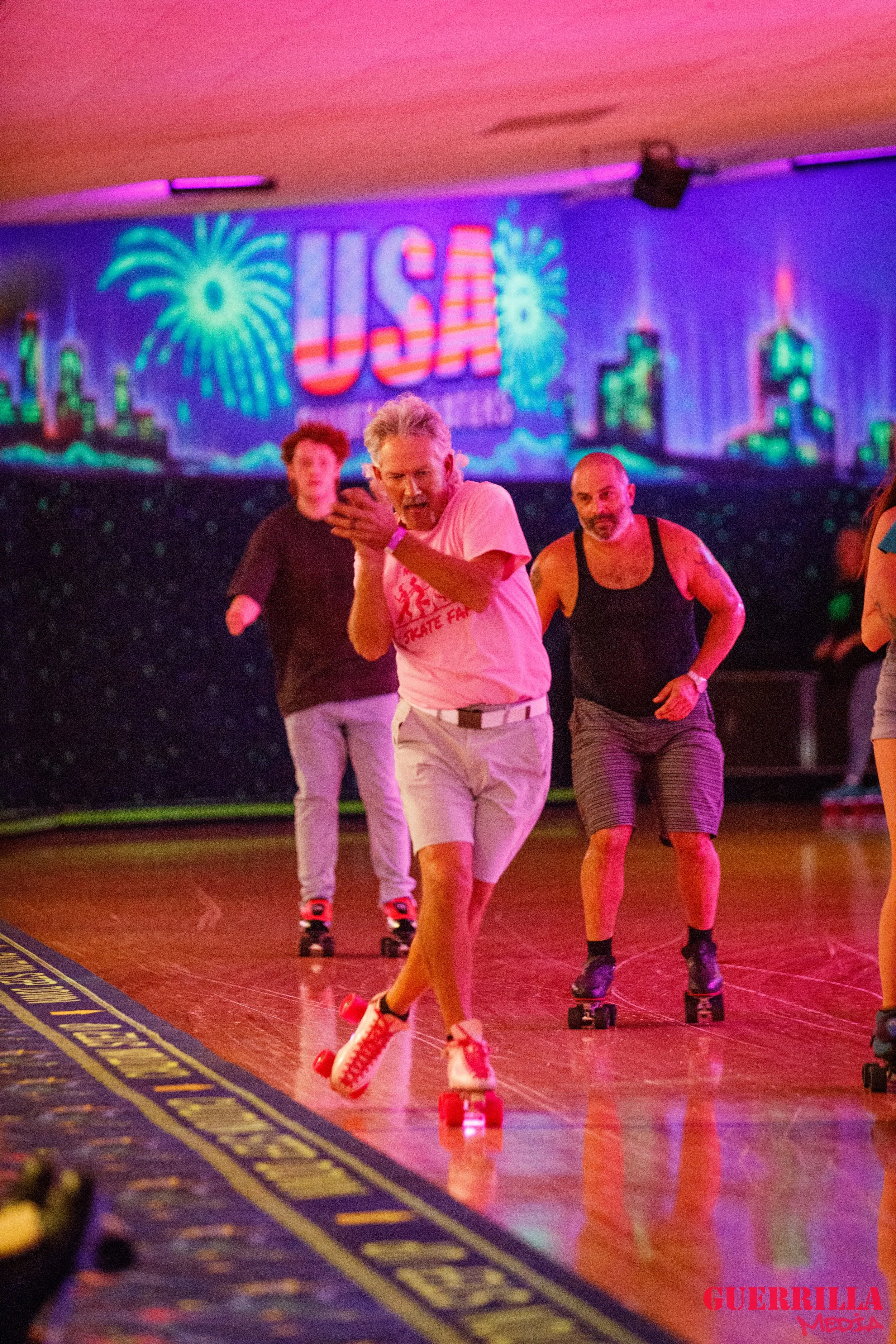 People roller skating in an indoor rink with a colorful background displaying fireworks and the word 'USA'.