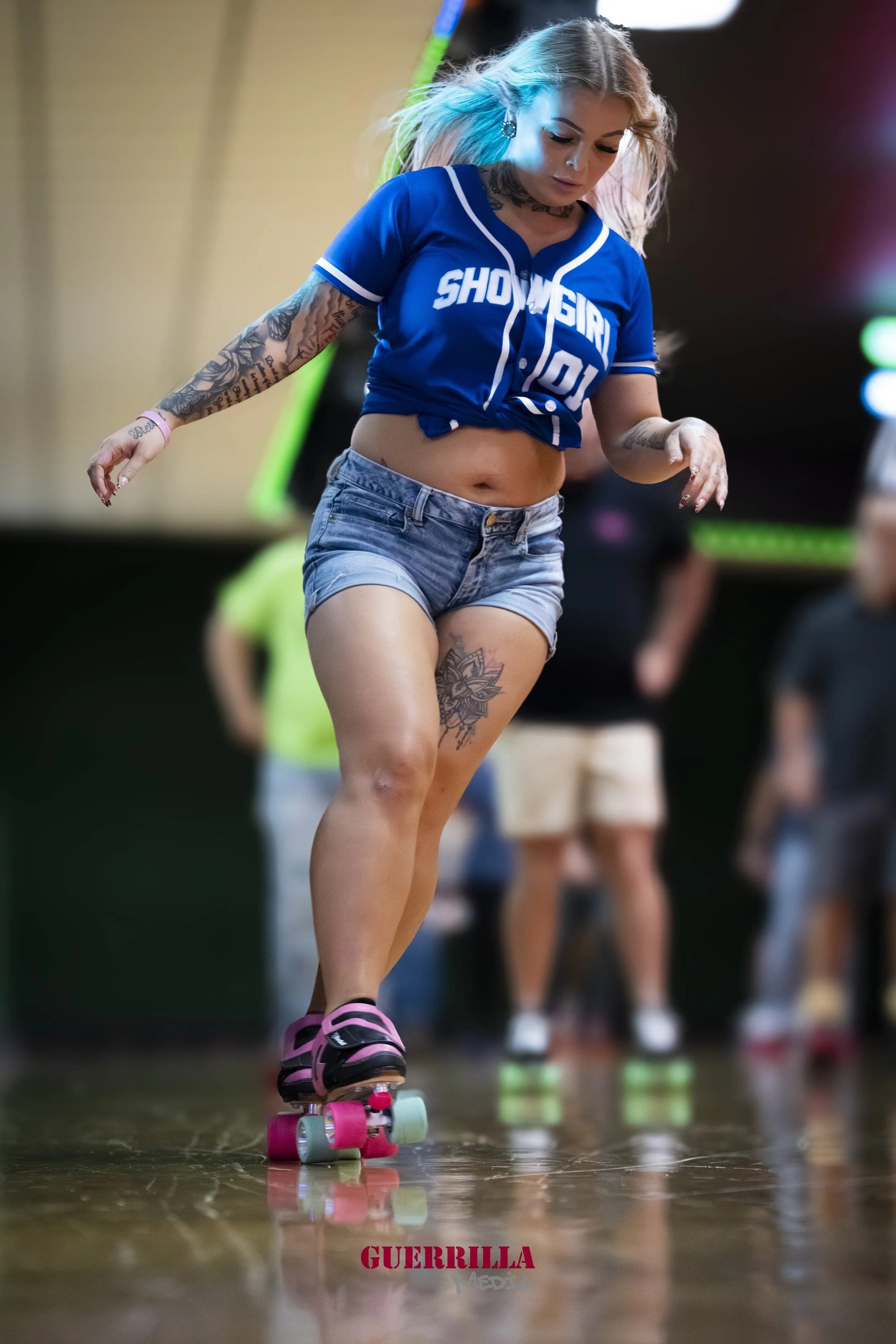 A young woman roller skating indoors, wearing a blue sports jersey with the words "SHOWGIRL 01", denim shorts, pink and black roller skates, and visible tattoos on her arms, thigh, and abdomen.