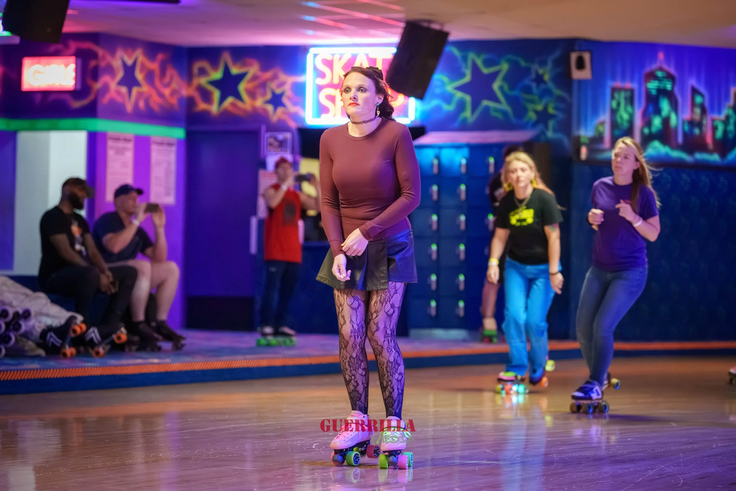 People roller skating in a neon-lit roller rink, with one woman in fishnet stockings and a brown long-sleeve top at the center, and others in the background skating. Some seated and watching, others skating on the floor.