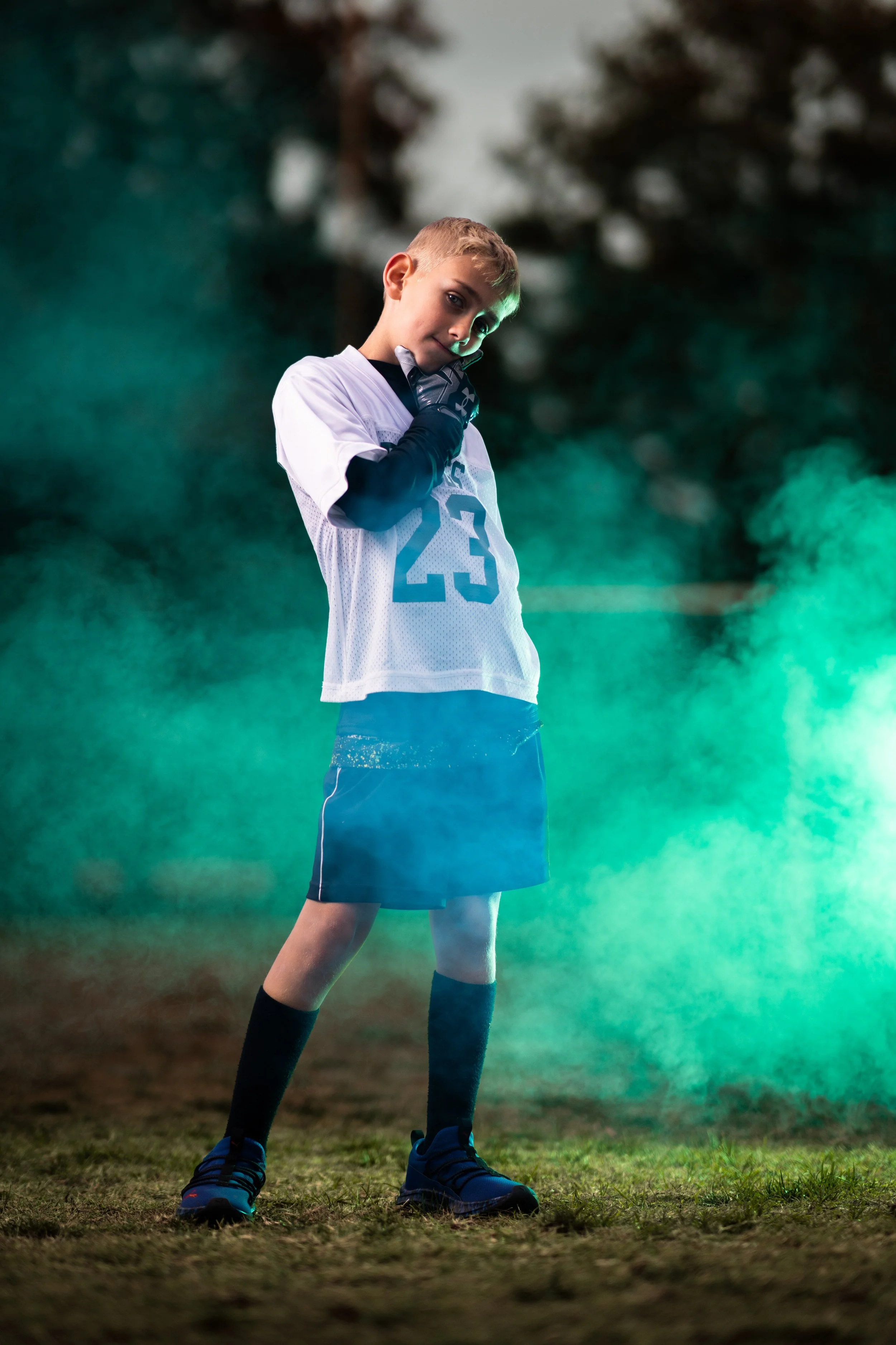 A young boy in a football uniform standing outdoors on grass at dusk, with a green light mist around him, wearing a white jersey with the number 23 and black shorts.