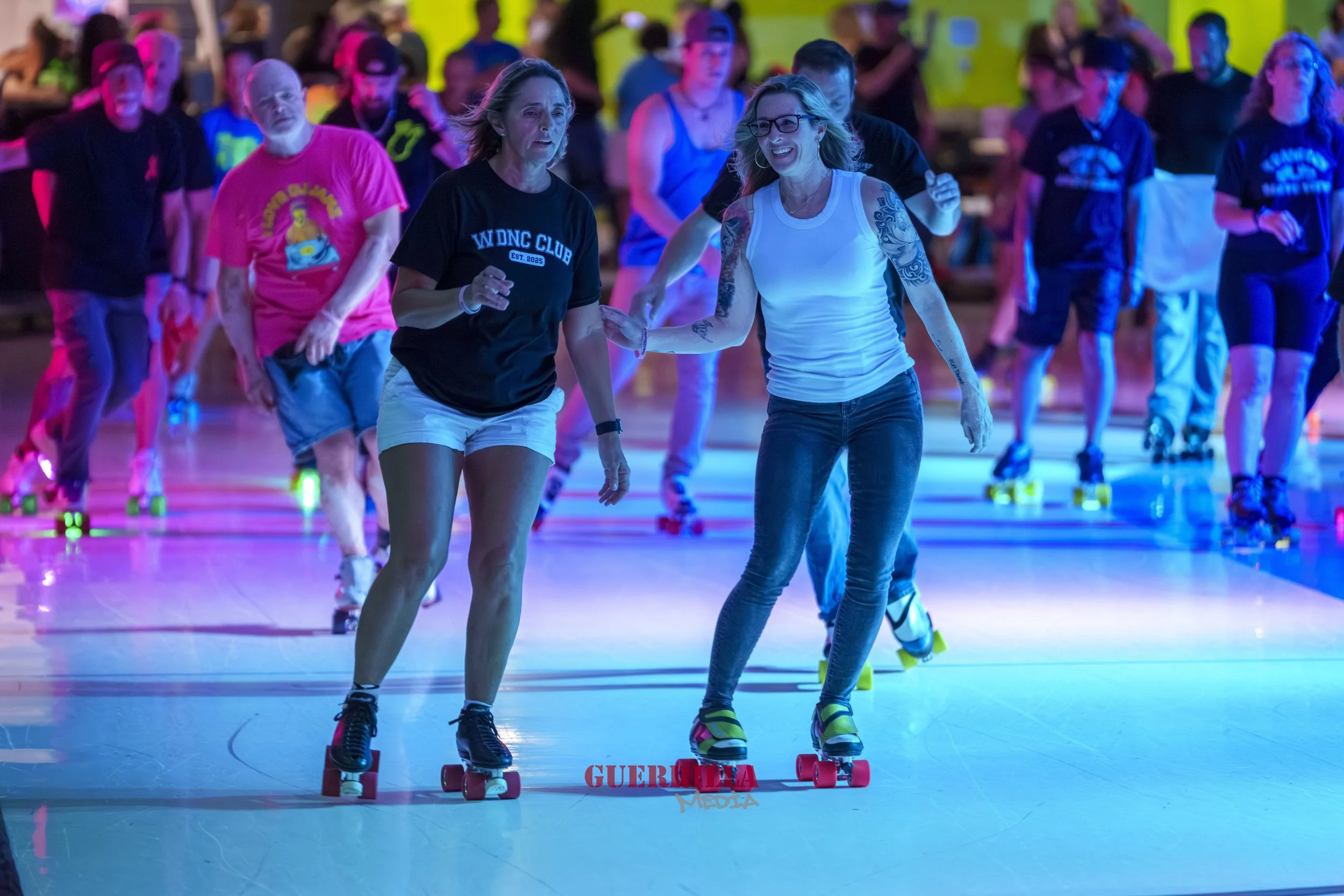 People roller skating together in a brightly lit indoor rink, with two women at the front, one assisting the other, smiling and enjoying the activity.