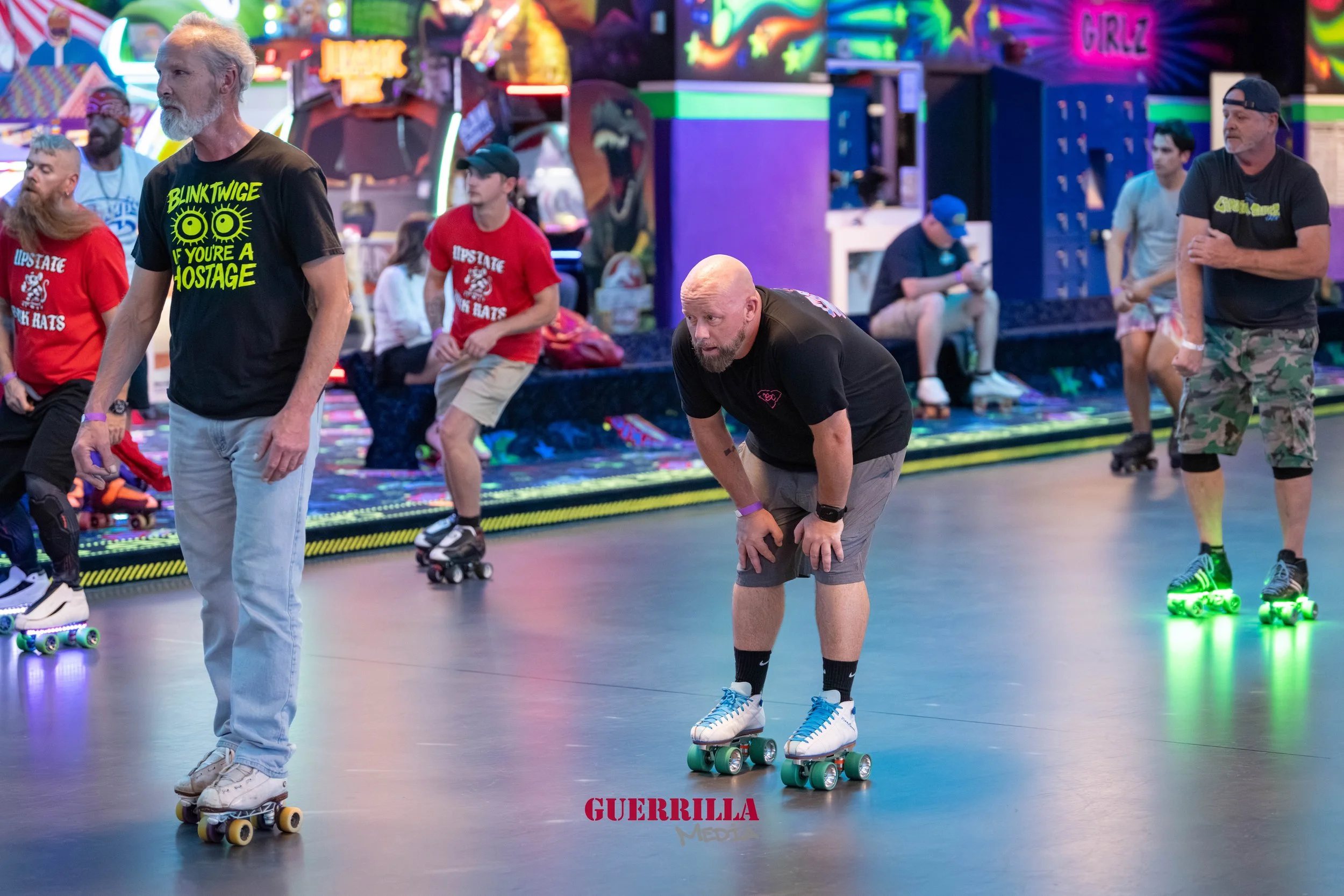 People roller skating in an indoor skating rink with colorful neon lights and arcade games in the background. The skaters are wearing casual clothes and appear focused or tired.