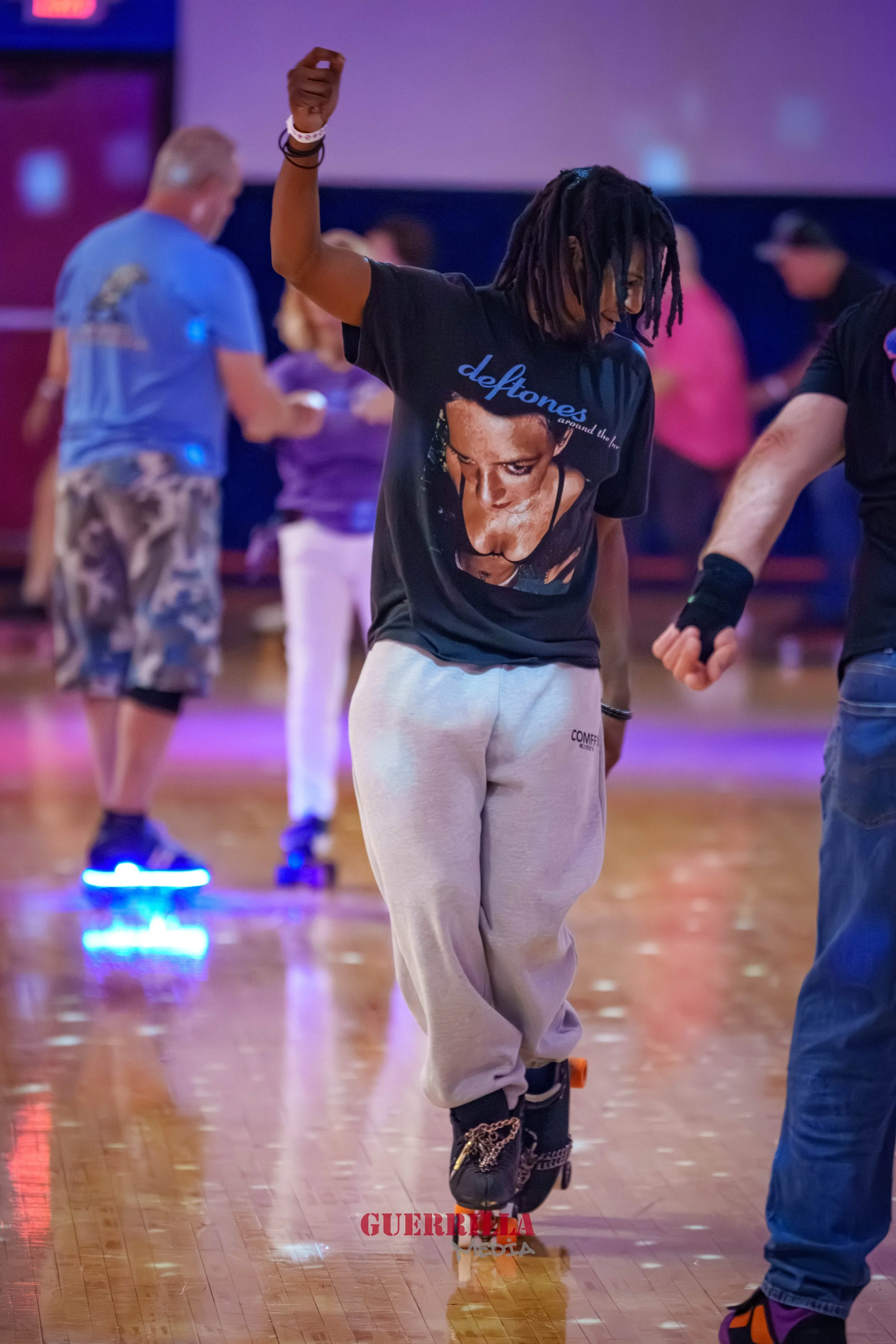 Person roller skating in a gym, wearing a Black T-shirt with a Michael Jackson print, grey sweatpants, and black skates, with several other people in the background.
