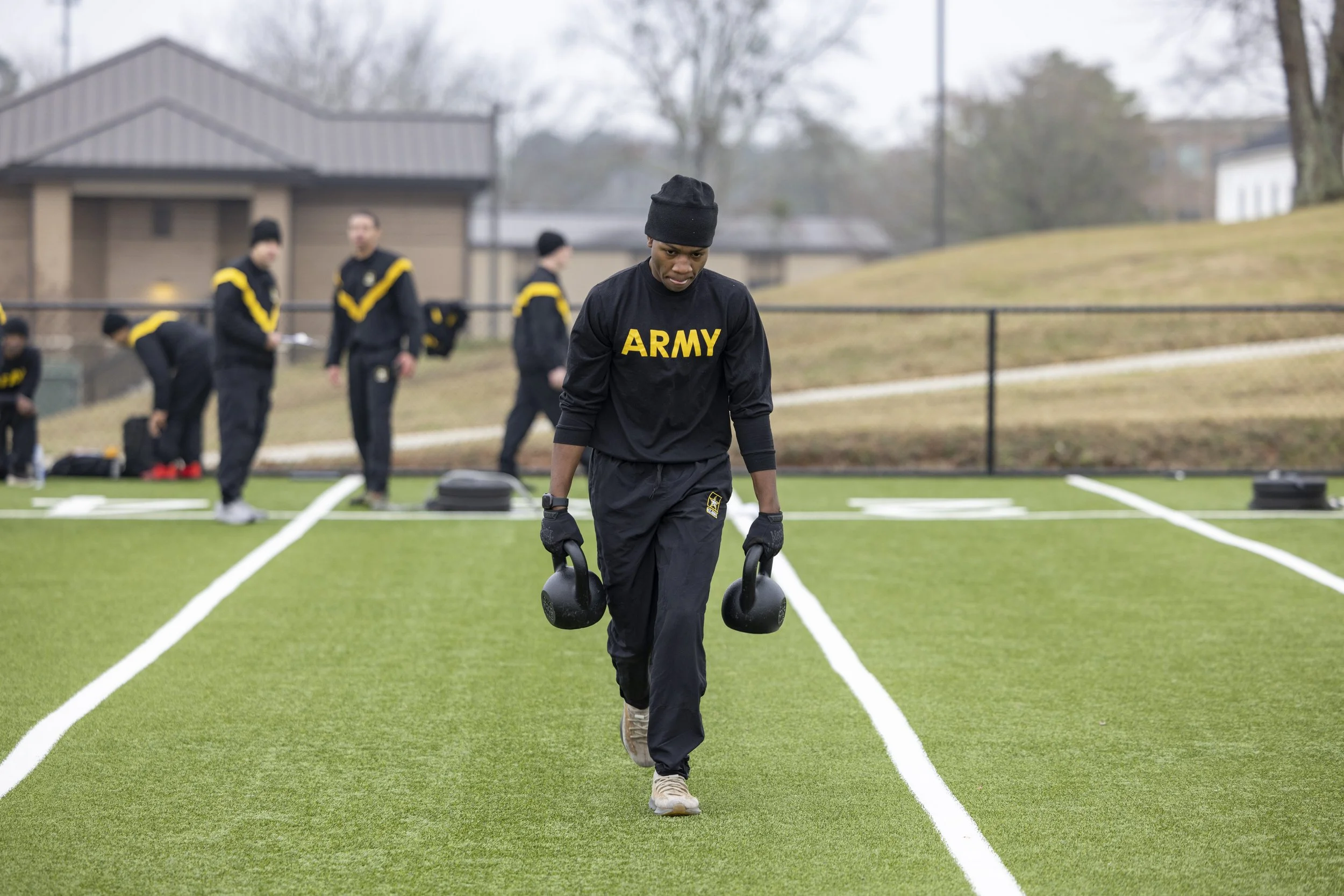 Military personnel in black uniforms with yellow accents exercises on a green field, with one person carrying kettlebells in the foreground and others in the background preparing for fitness training.