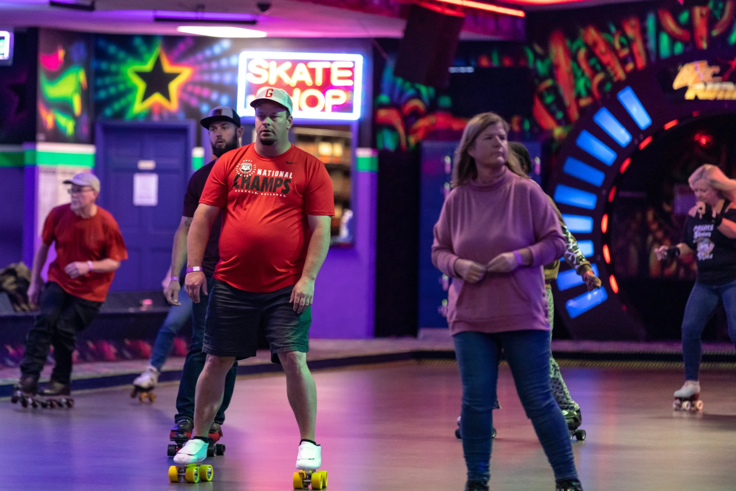 People roller skating in an indoor roller rink with neon lights and colorful wall decorations, including a sign that reads 'Skate Shop'.