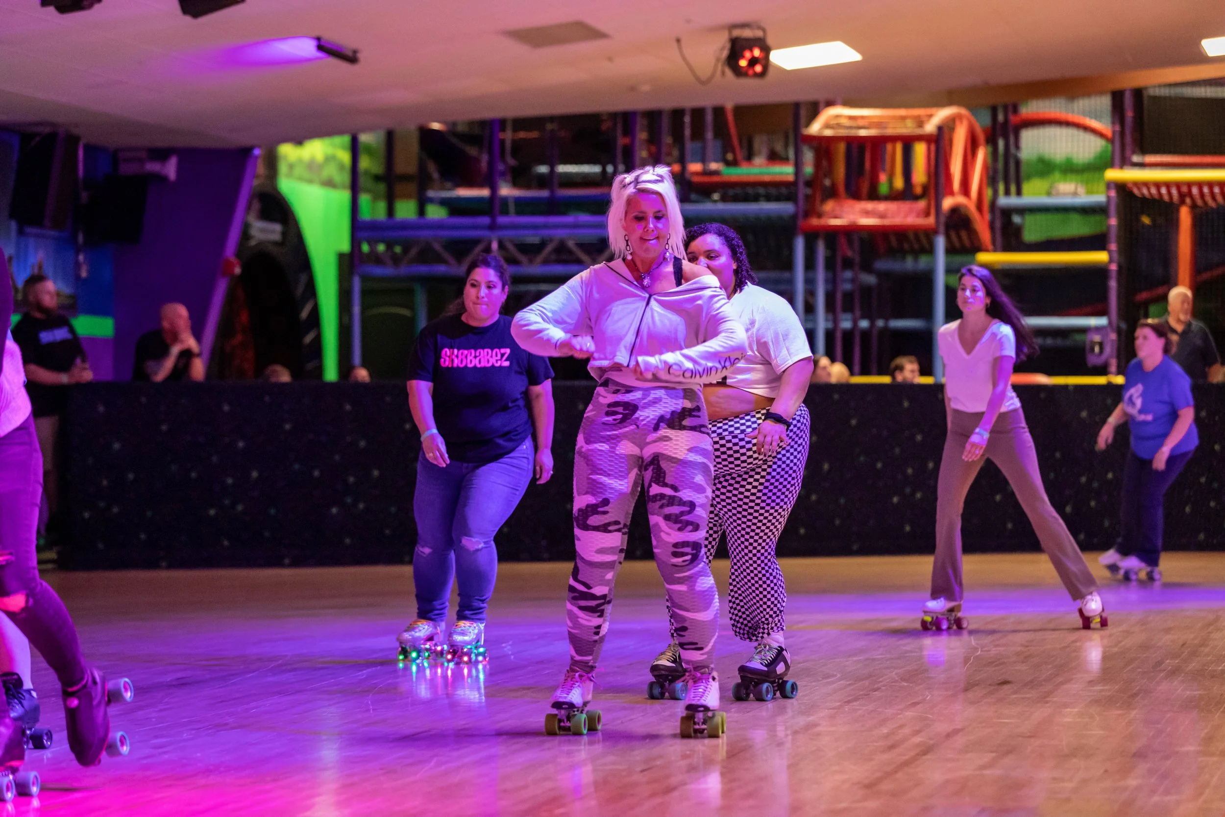 People roller skating inside an indoor skating rink with colorful lighting and play structures in the background.