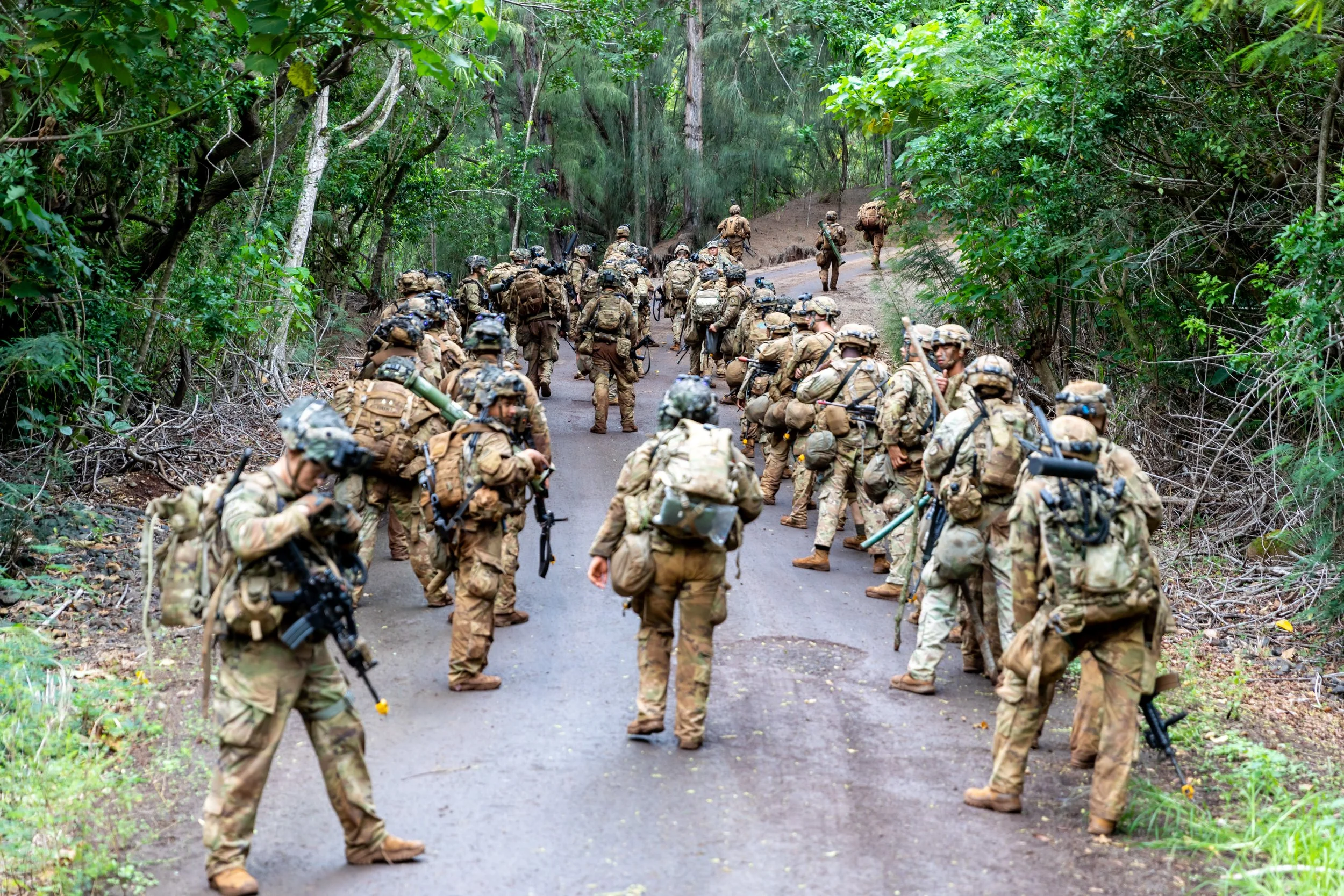 A group of soldiers in camouflage uniforms and gear walking on a forested dirt road.