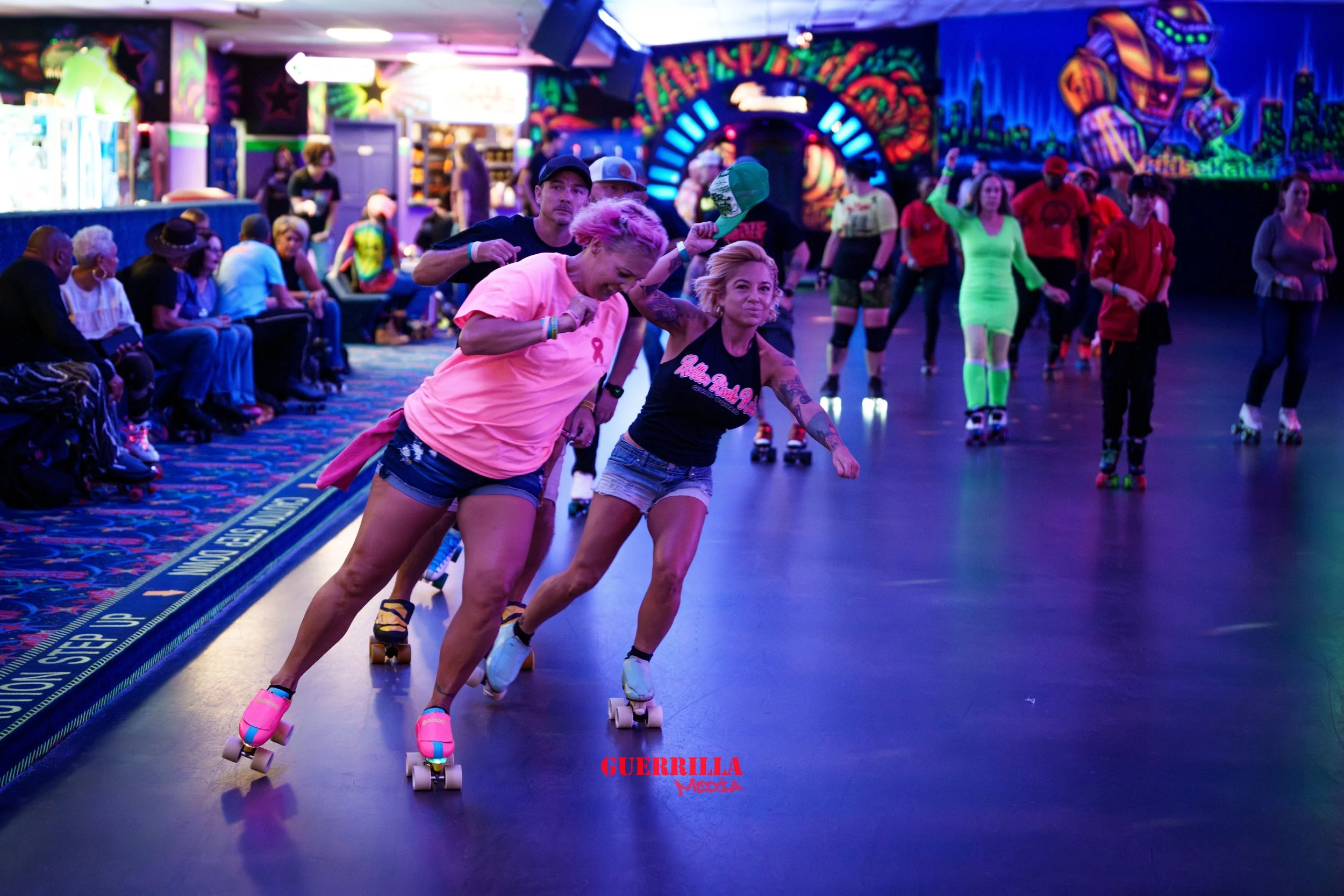 People roller skating in an indoor roller rink with colorful neon lights and graffiti-style wall art, some seated and watching, others skating or standing.