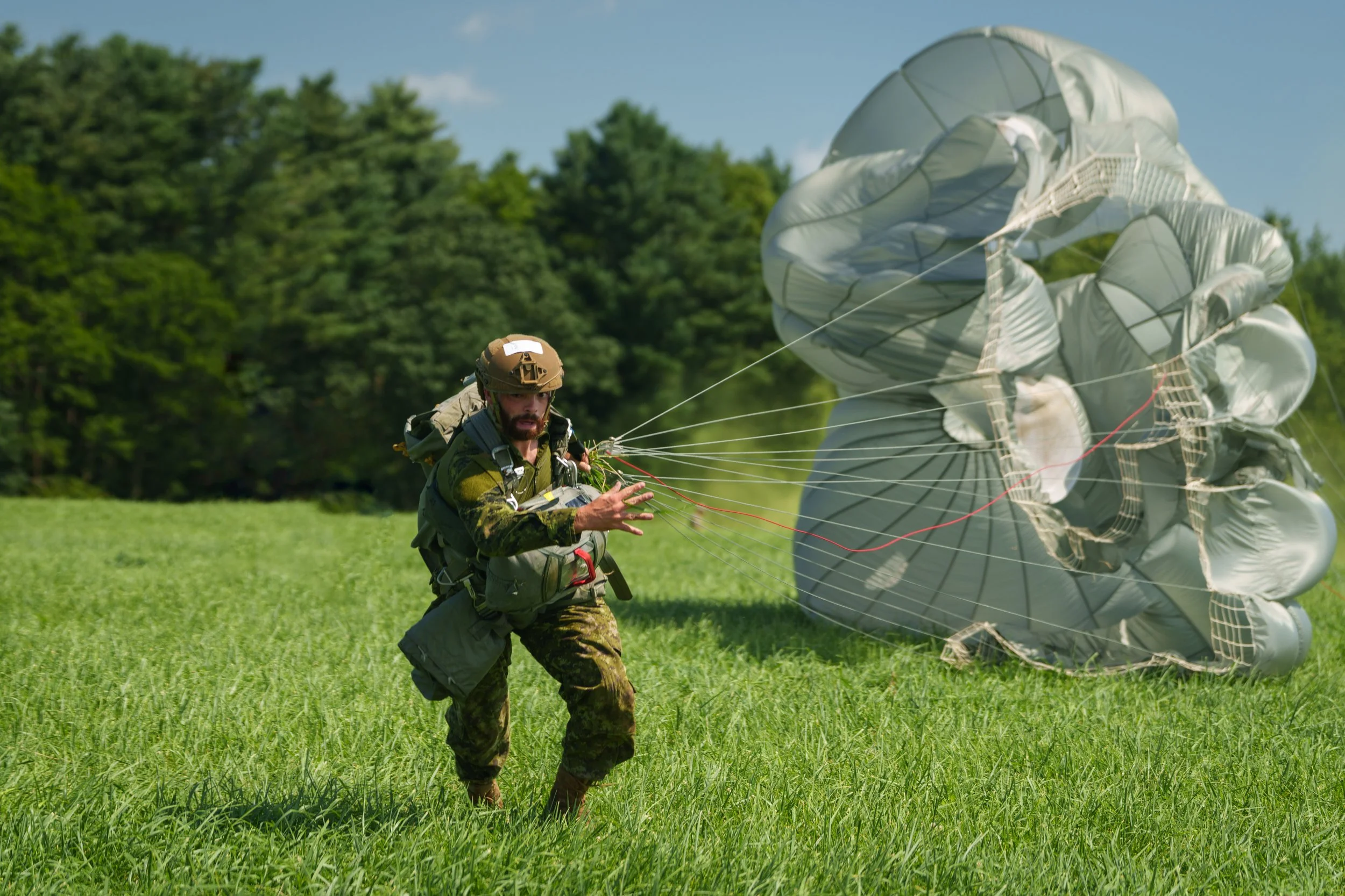 A soldier on a grassy field prepares for a parachute jump, with a large parachute deployed in the background.