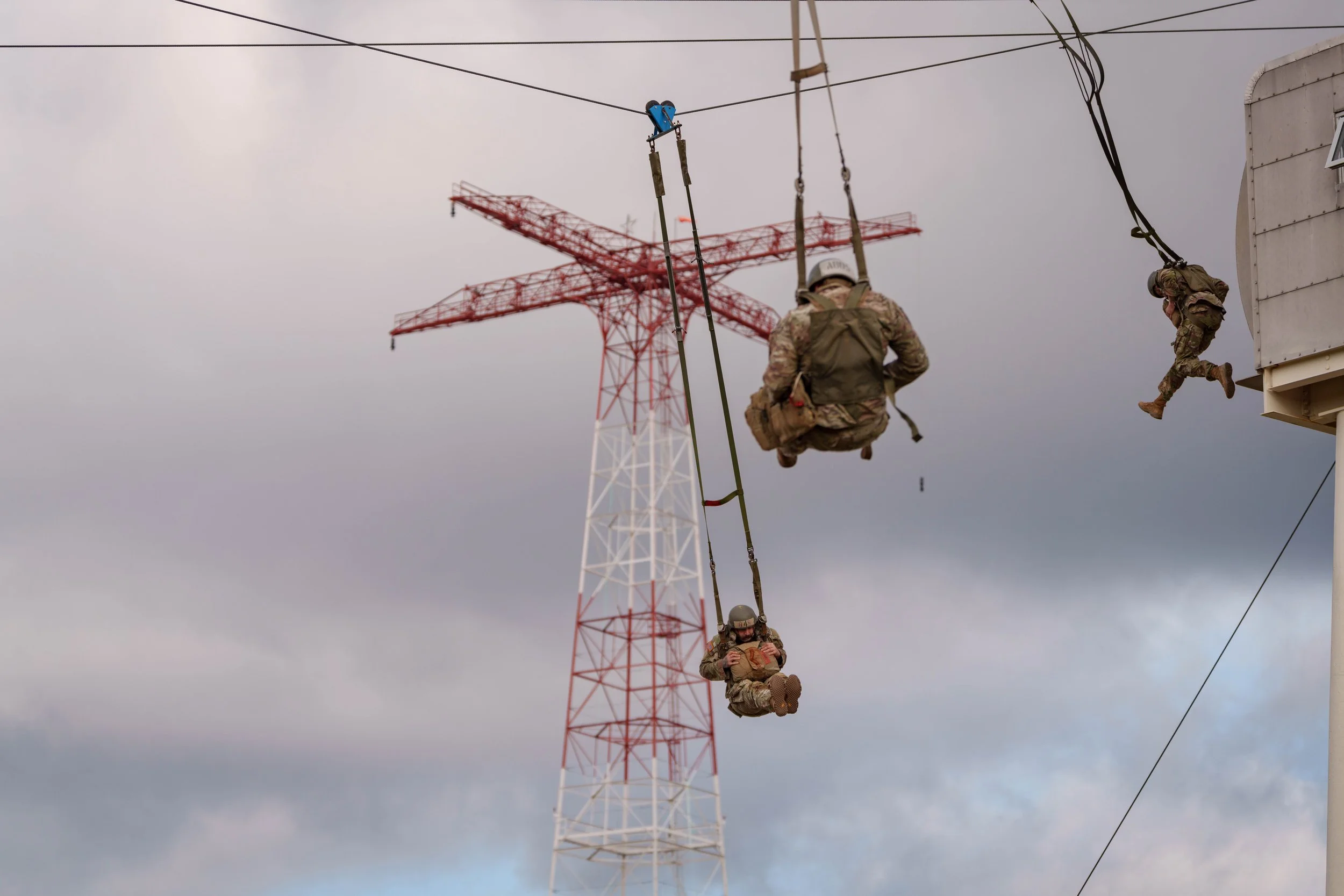 Three soldiers, in military gear, swinging on a cable from a building with a large red and white transmission tower in the background on a cloudy day.