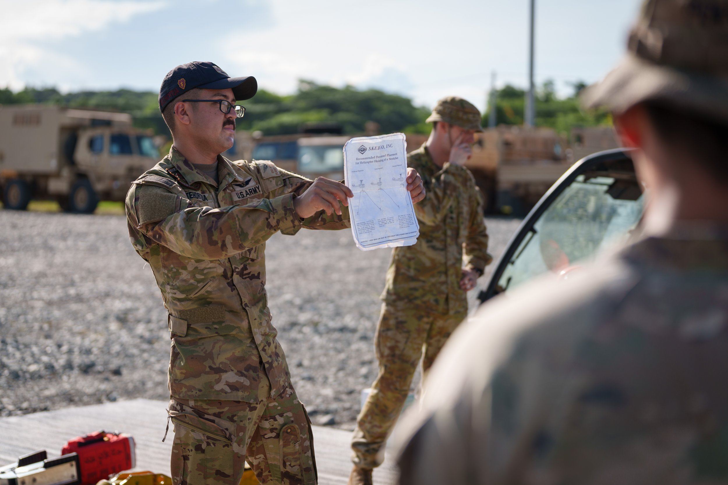 Military personnel in camouflage uniforms standing outdoors, one holding a diagram or map, with military vehicles in the background.